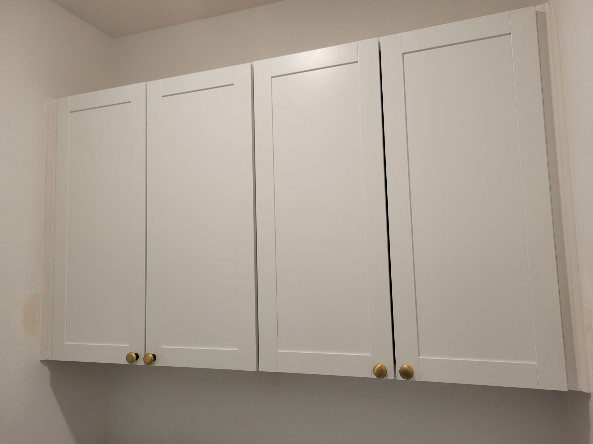 Four white kitchen cabinets with gold knobs mounted on a light-colored wall.