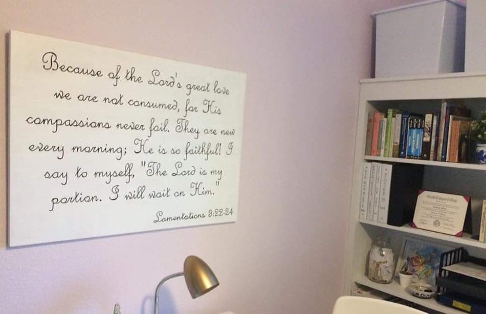 A room corner with a scripture sign on a wall, a bookshelf, and a desk lamp.