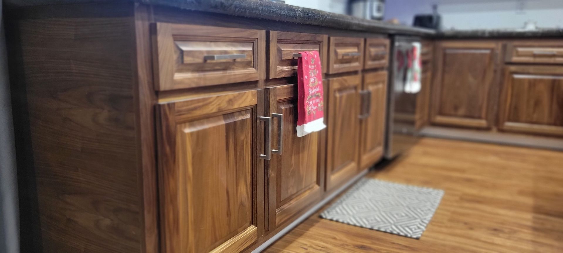Kitchen cabinets with a wooden finish, a red towel hanging on the front, and a gray rug on the floor.