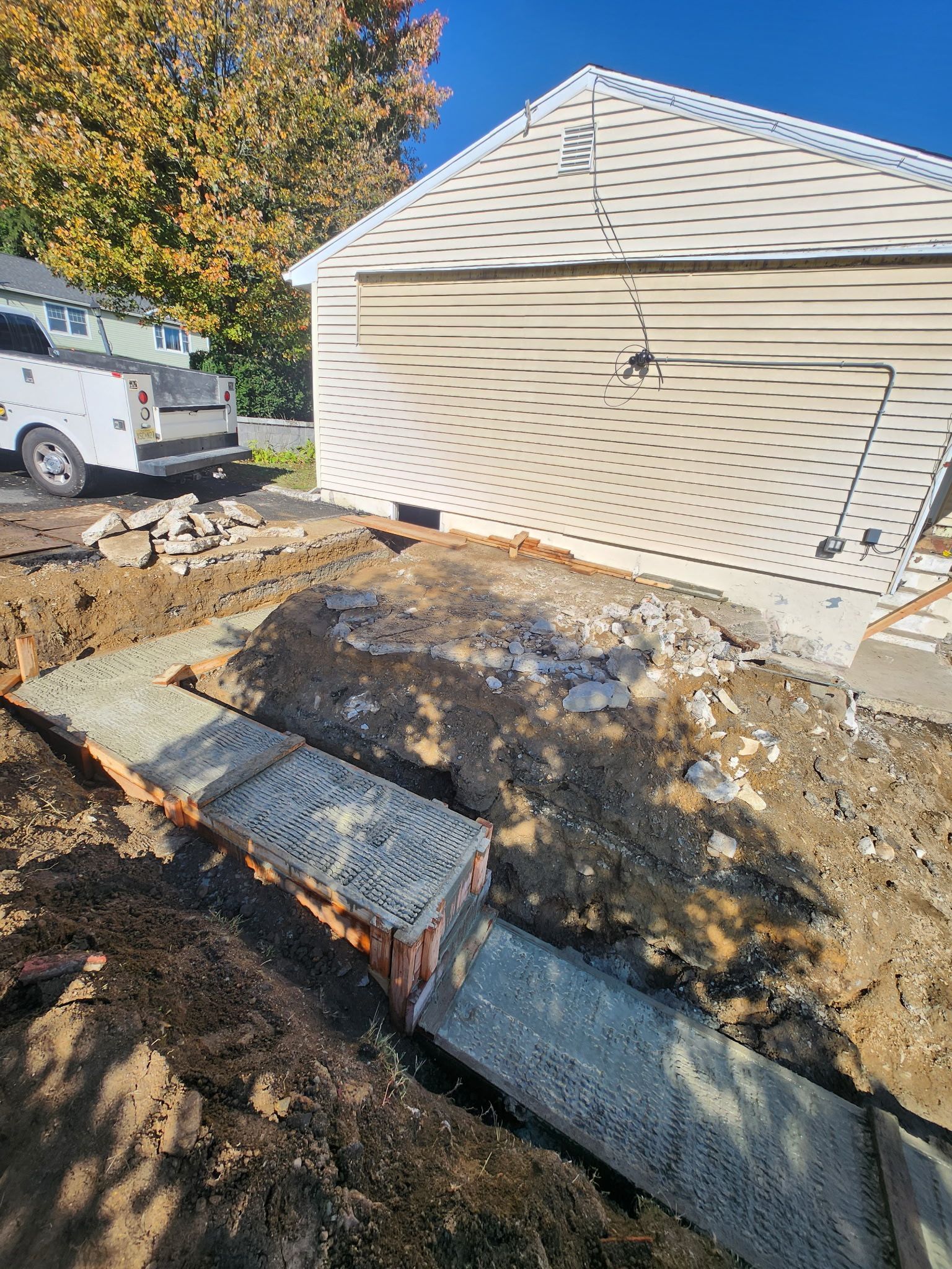 Backyard excavation beside a beige garage, with a white van and exposed drain pipes and dirt.