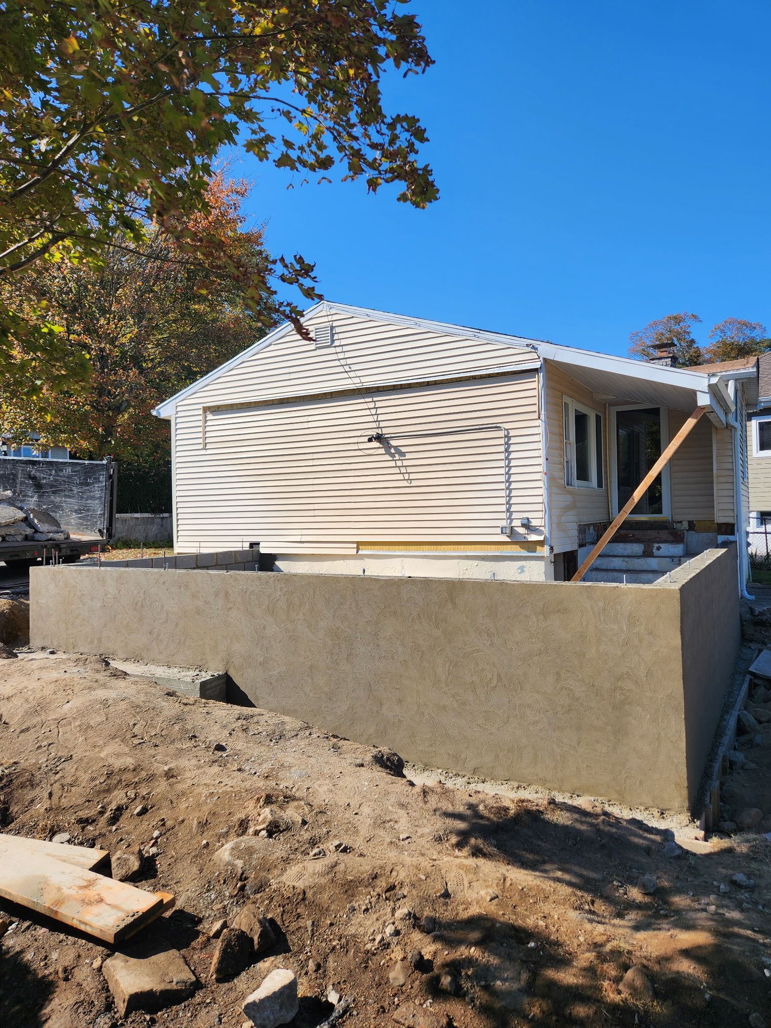 House with beige siding behind a tall concrete retaining wall on a dirt slope under a blue sky