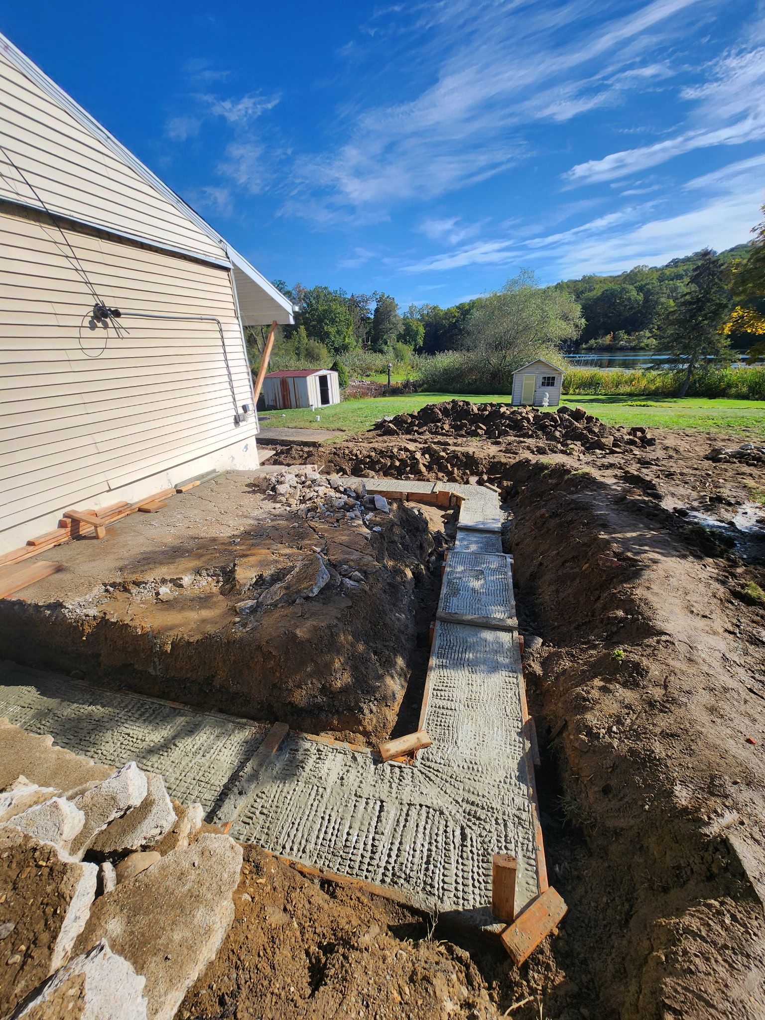 Backyard excavation beside a house, with a concrete footing trench and blue sky overhead