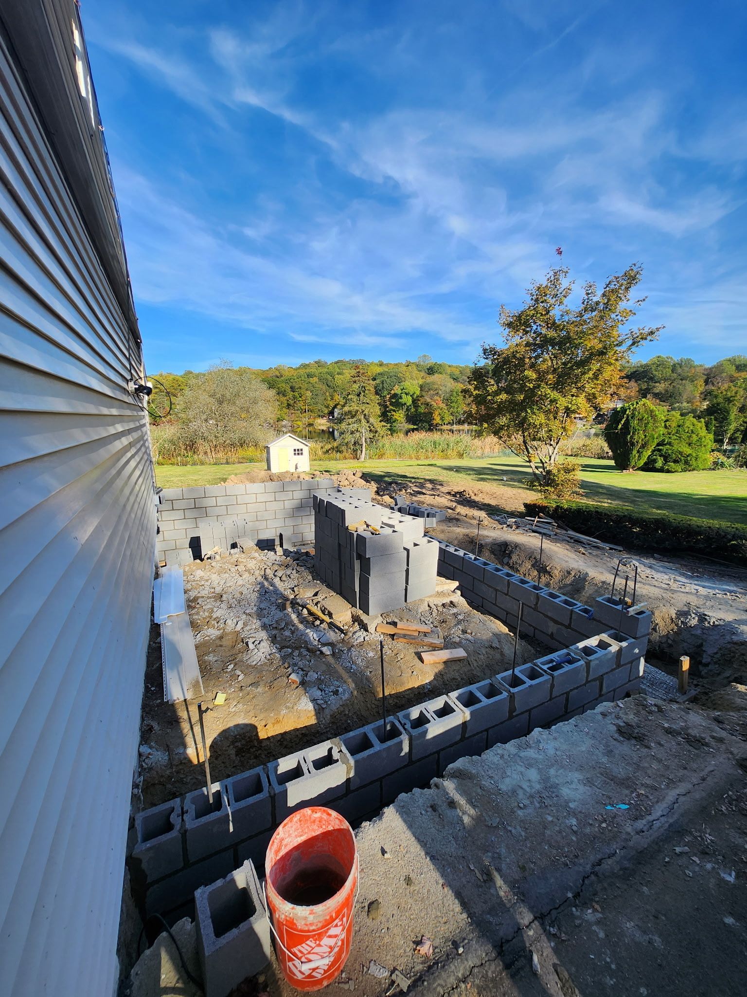 Construction site foundation with cinder blocks, a red bucket, and a house exterior under a blue sky