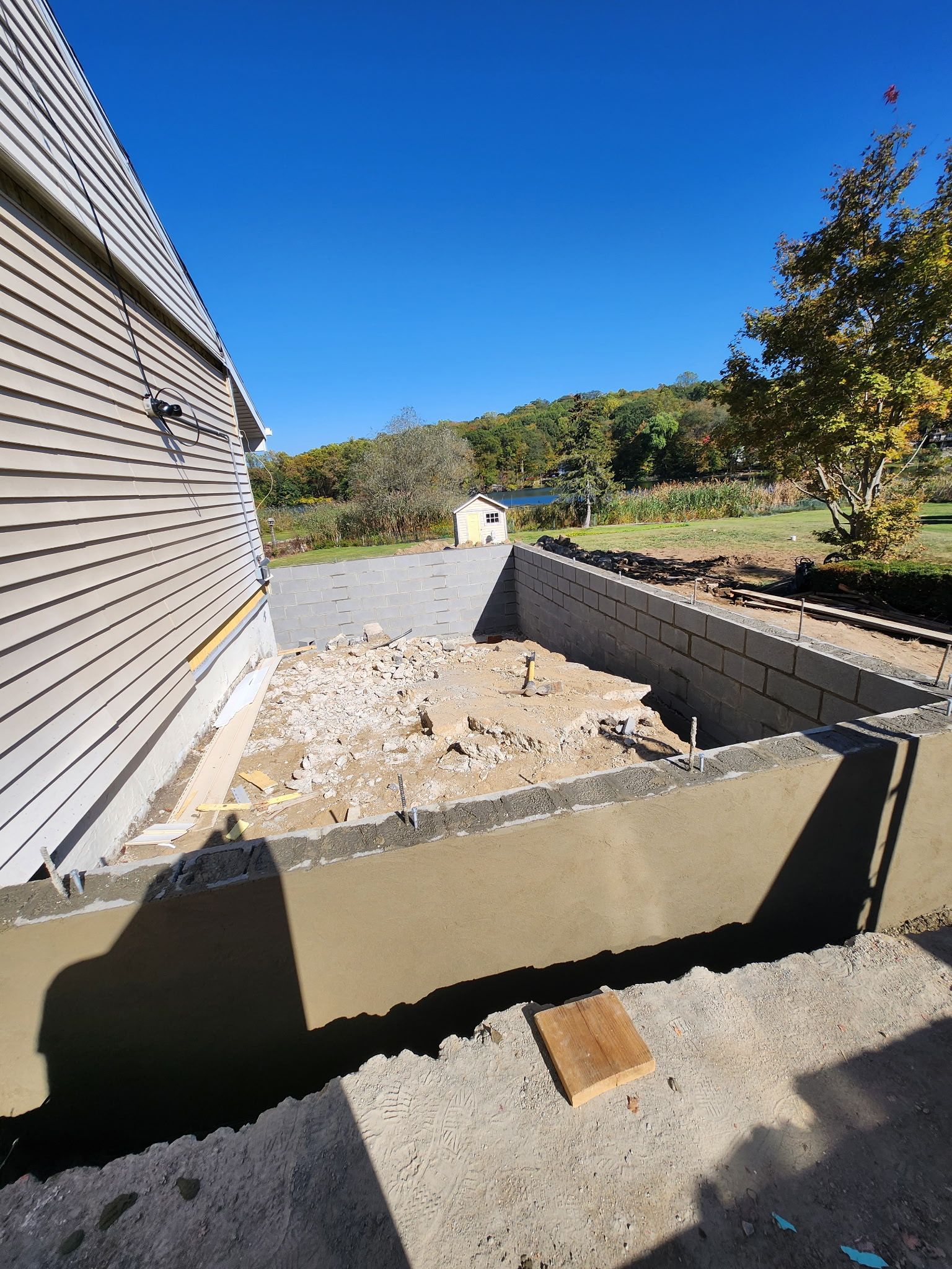 Empty concrete foundation beside a gray-sided house, with rubble and a sunny hillside view beyond
