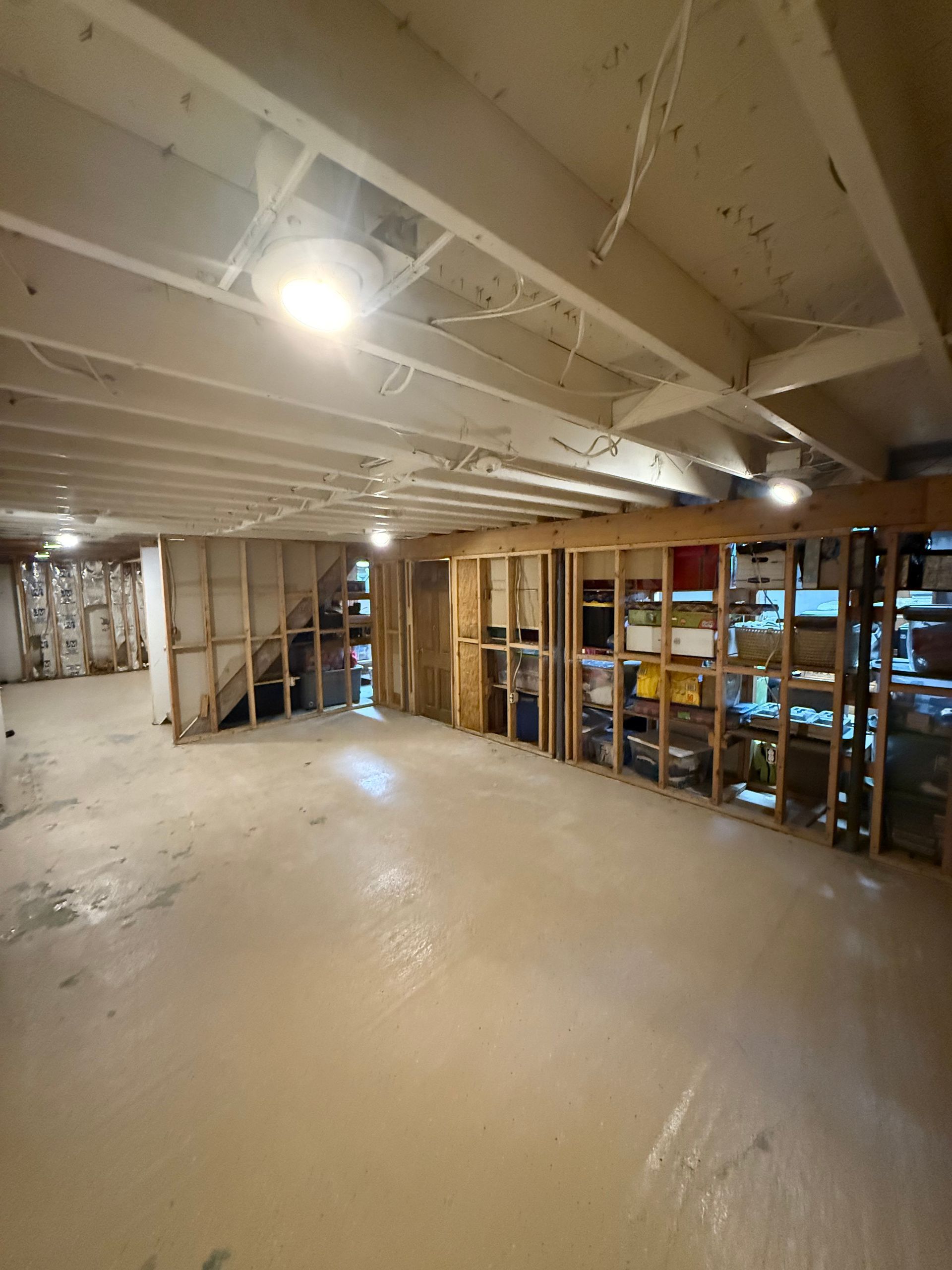 Renovated basement with exposed beams, bare floor, framed wall on right, and bright ceiling light.