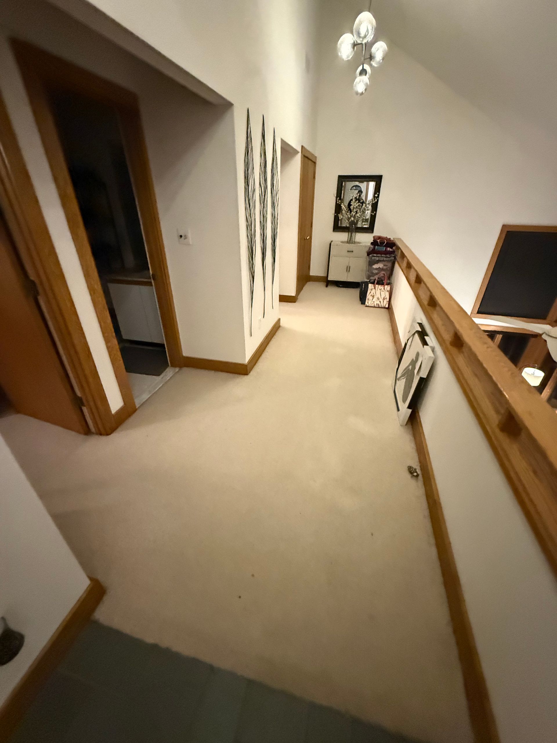 Hallway with beige carpet, white walls, railing, and stairs leading to a small landing with a chair and wall art