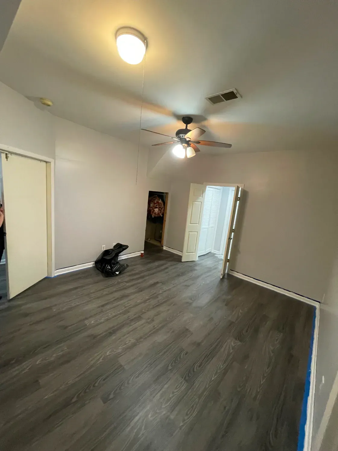 Empty room with grey wood-look flooring, pale walls, open doors, and ceiling fan. A black trash bag sits in the middle.