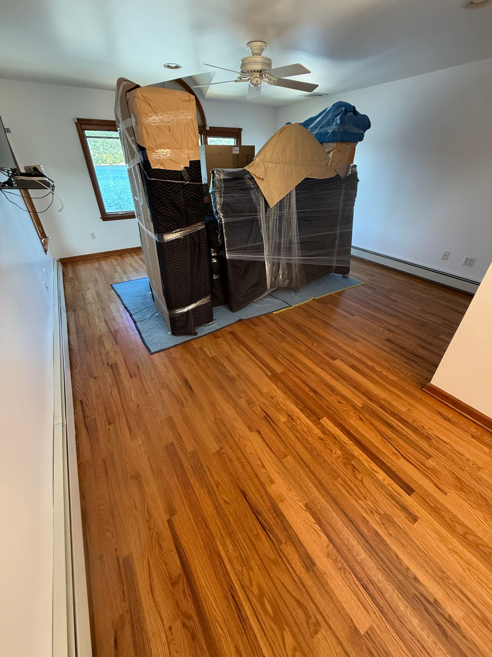 Empty room with hardwood floors and cardboard-wrapped furniture near the back wall