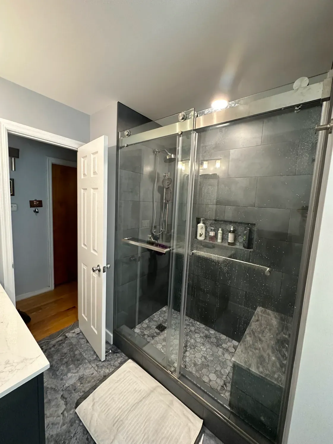 Bathroom with glass shower, dark gray tiled walls, bench, open door to bedroom, gray floor.