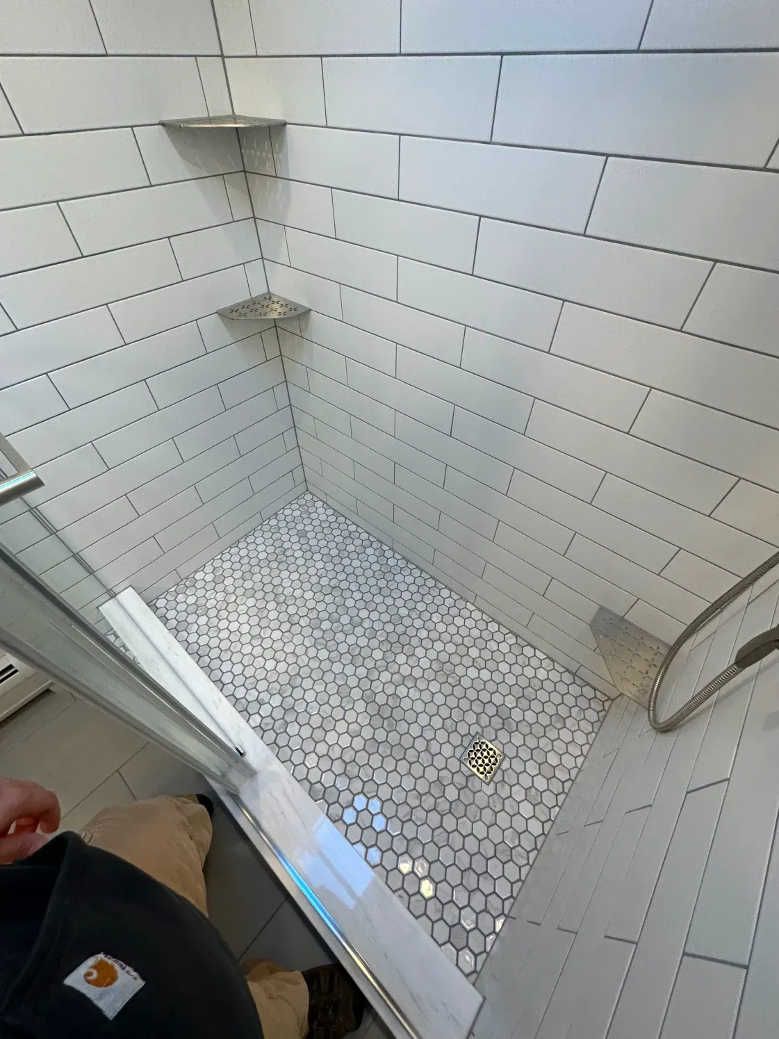 White tiled shower with glass door, penny tile floor, and two corner shelves.
