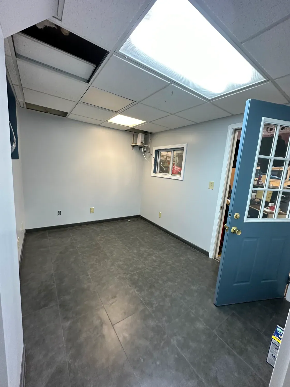 Empty office room with gray walls, floor, and a blue door. Ceiling tiles and window visible.
