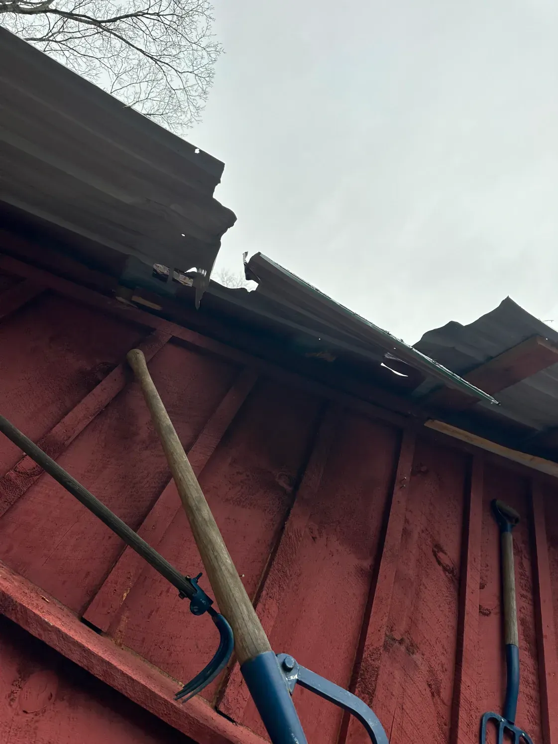 Red shed with damaged corrugated metal roof, tools hanging on the wall, cloudy sky.