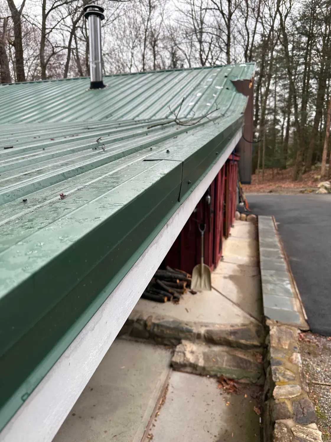 Green metal roof of a building with chimney. Red siding, concrete steps, and a black paved area are visible.