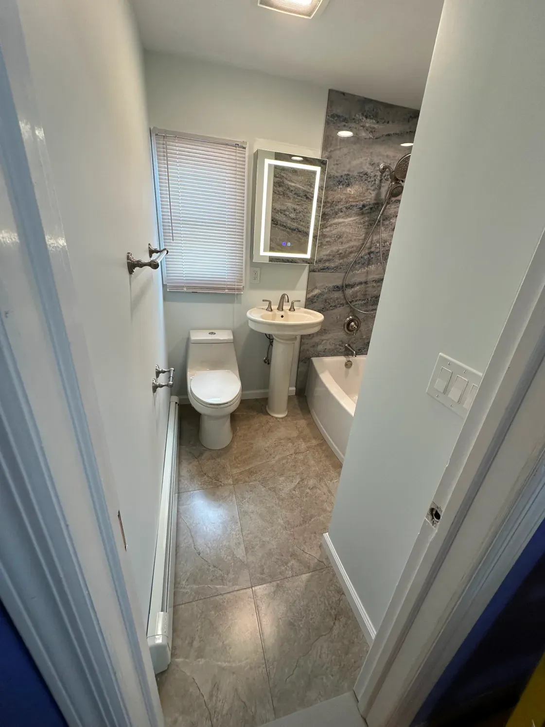 Narrow bathroom with gray tile, white toilet, pedestal sink, and marble shower wall.