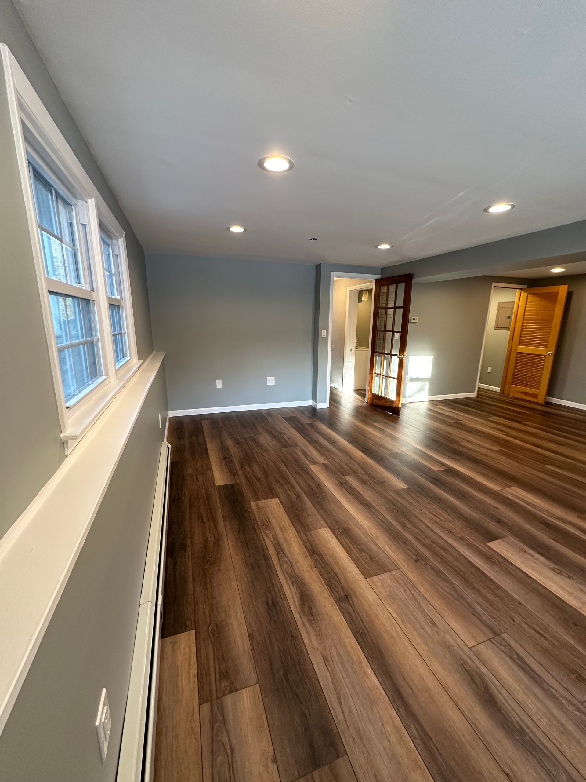 Empty living room with gray walls, wood floors, recessed lights, and large windows on the left.