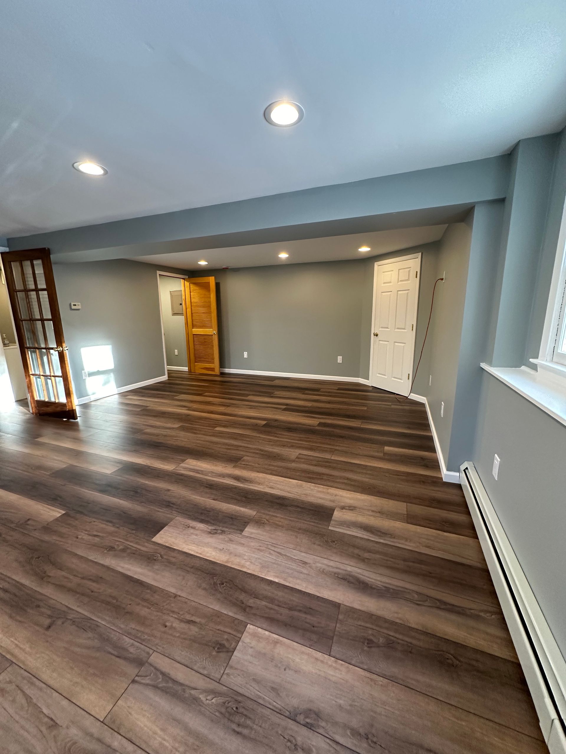Empty living room with wood flooring, gray walls, recessed lights, and a white door near windows.