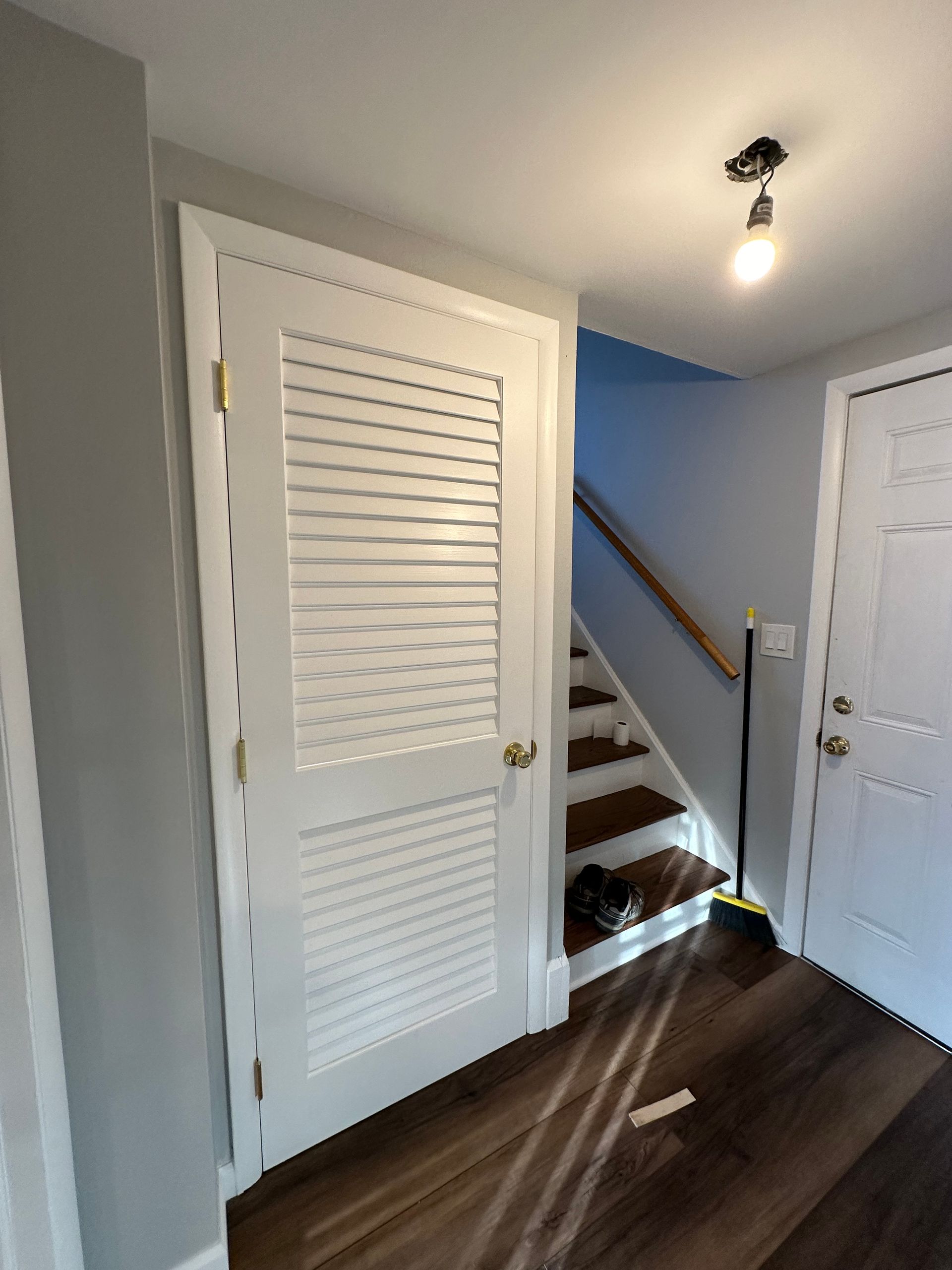 White hallway with louvered closet doors, stairs, and dark wood floor under a ceiling light.