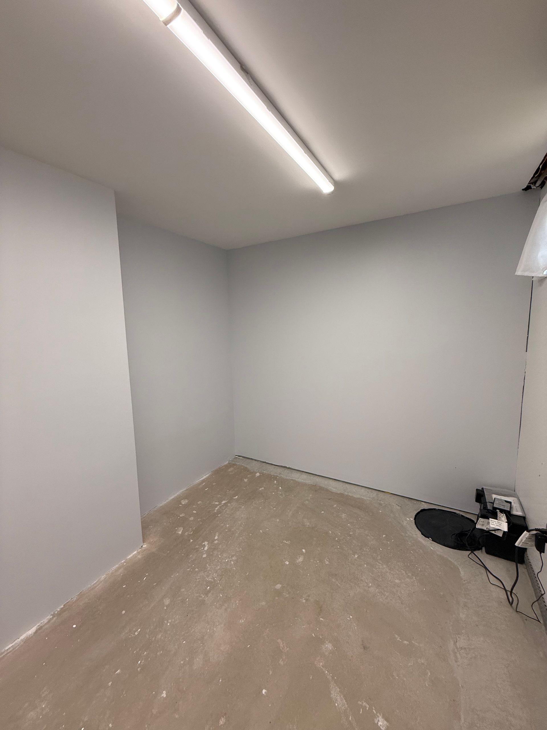 Empty beige room with white walls, fluorescent ceiling light, and a black item on the floor in the corner
