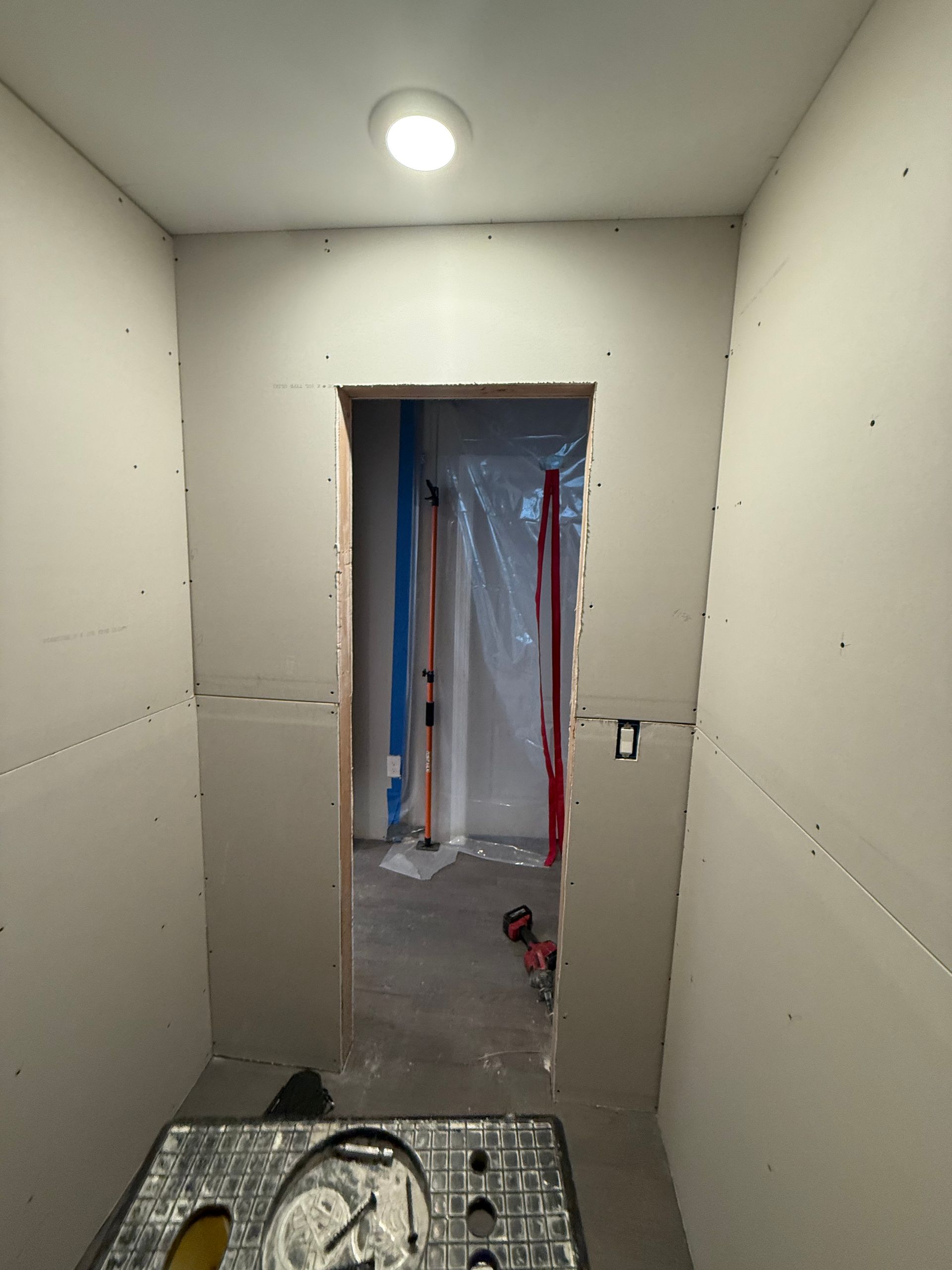 Small unfinished hallway with drywall, a lit ceiling bulb, and a doorway leading into a work area.