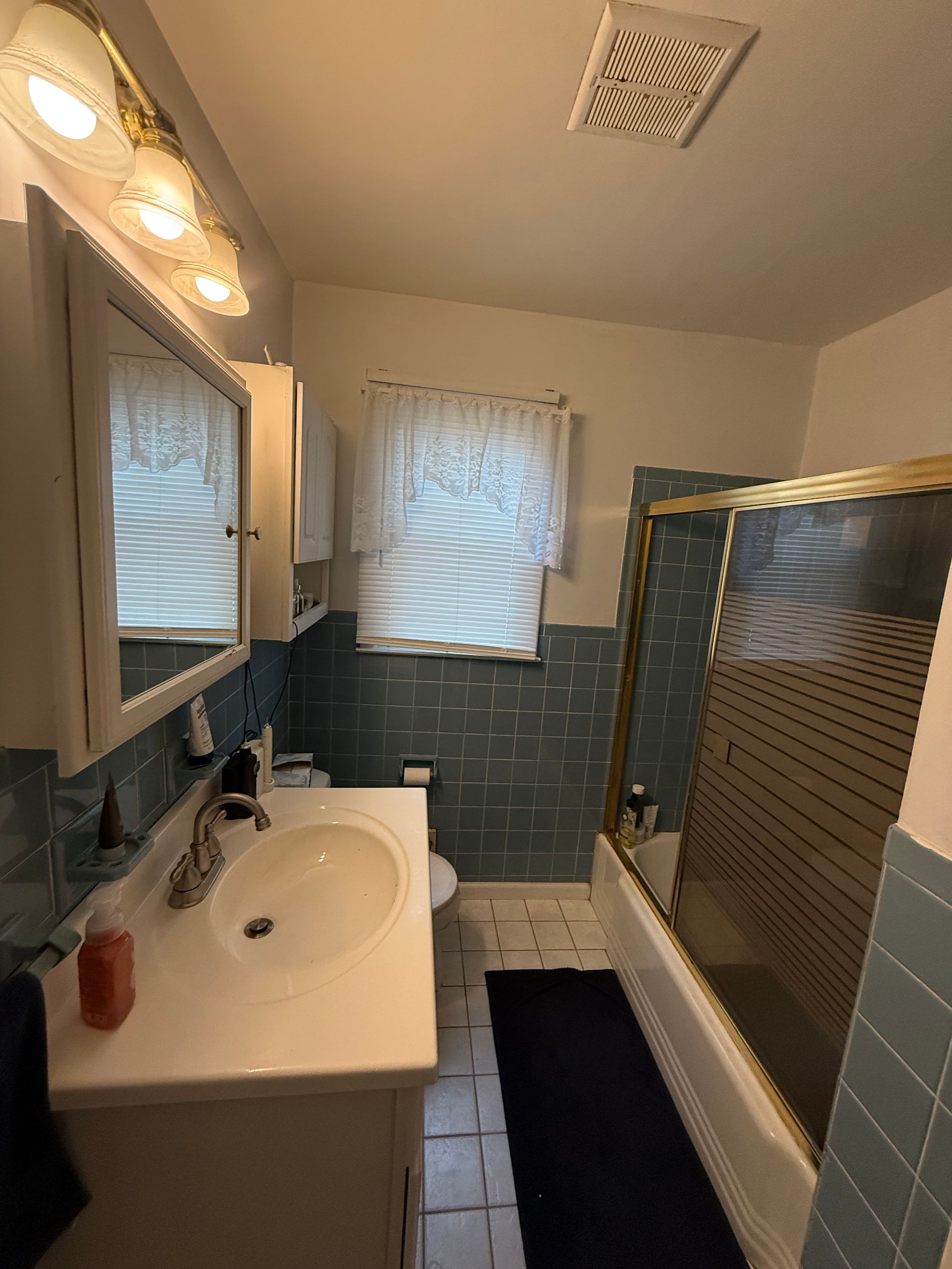 Small bathroom with white sink, blue tile shower, vanity lights, and window blinds.
