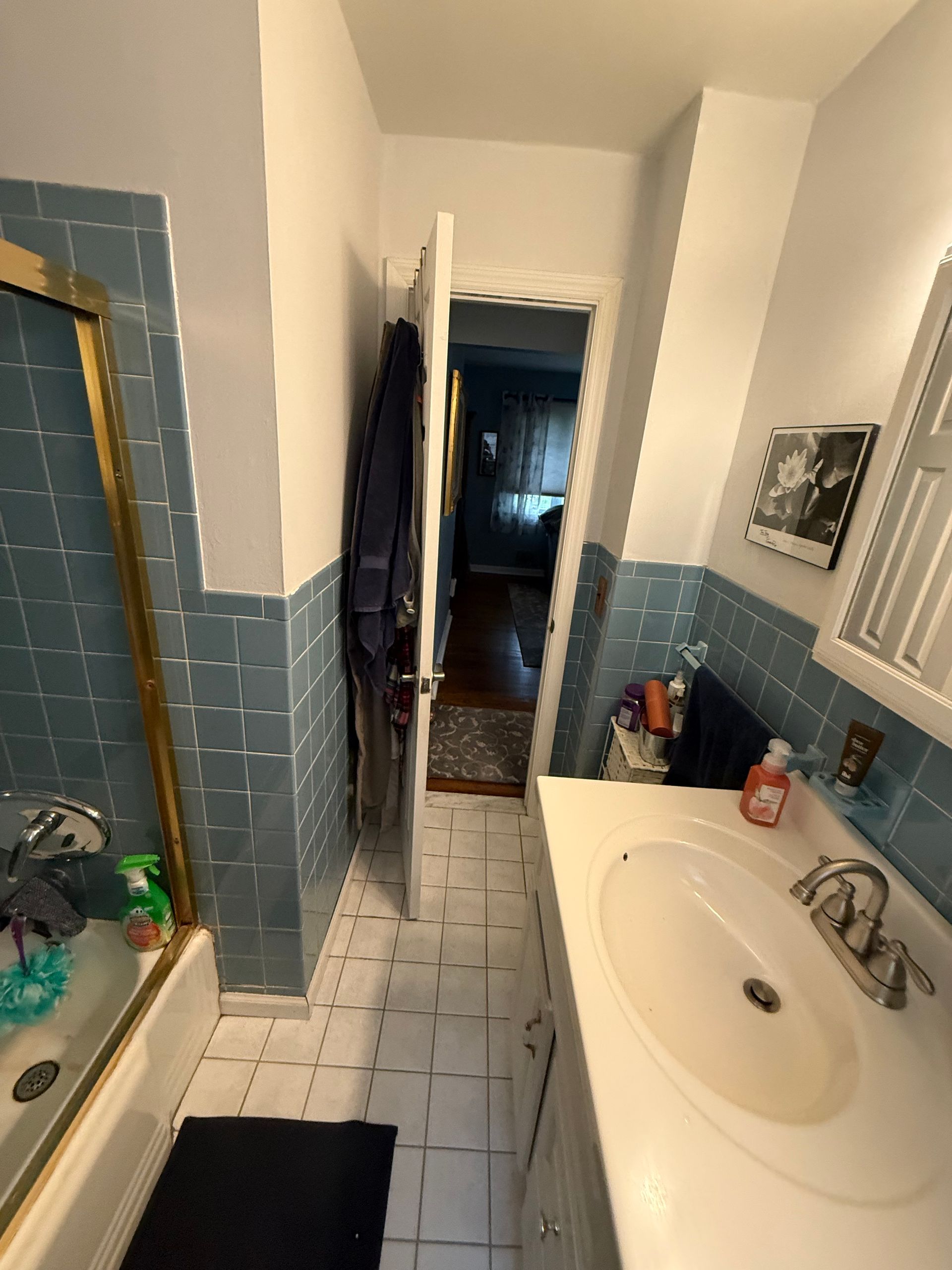 Narrow bathroom with blue tile, white sink, mirror, bathtub, and an open door to a dark hallway.