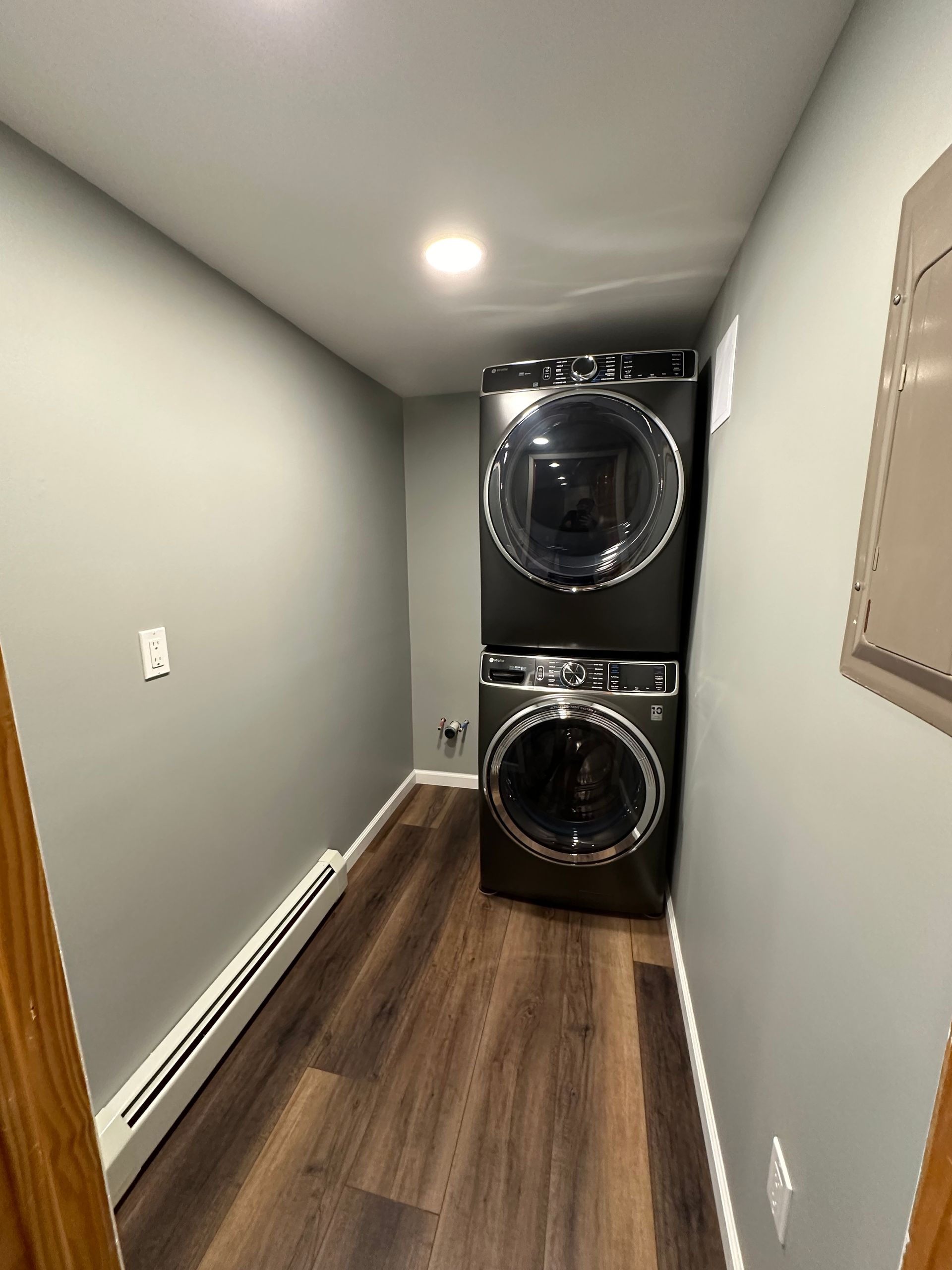 Laundry room with stacked black washer and dryer; gray walls, wood floor.