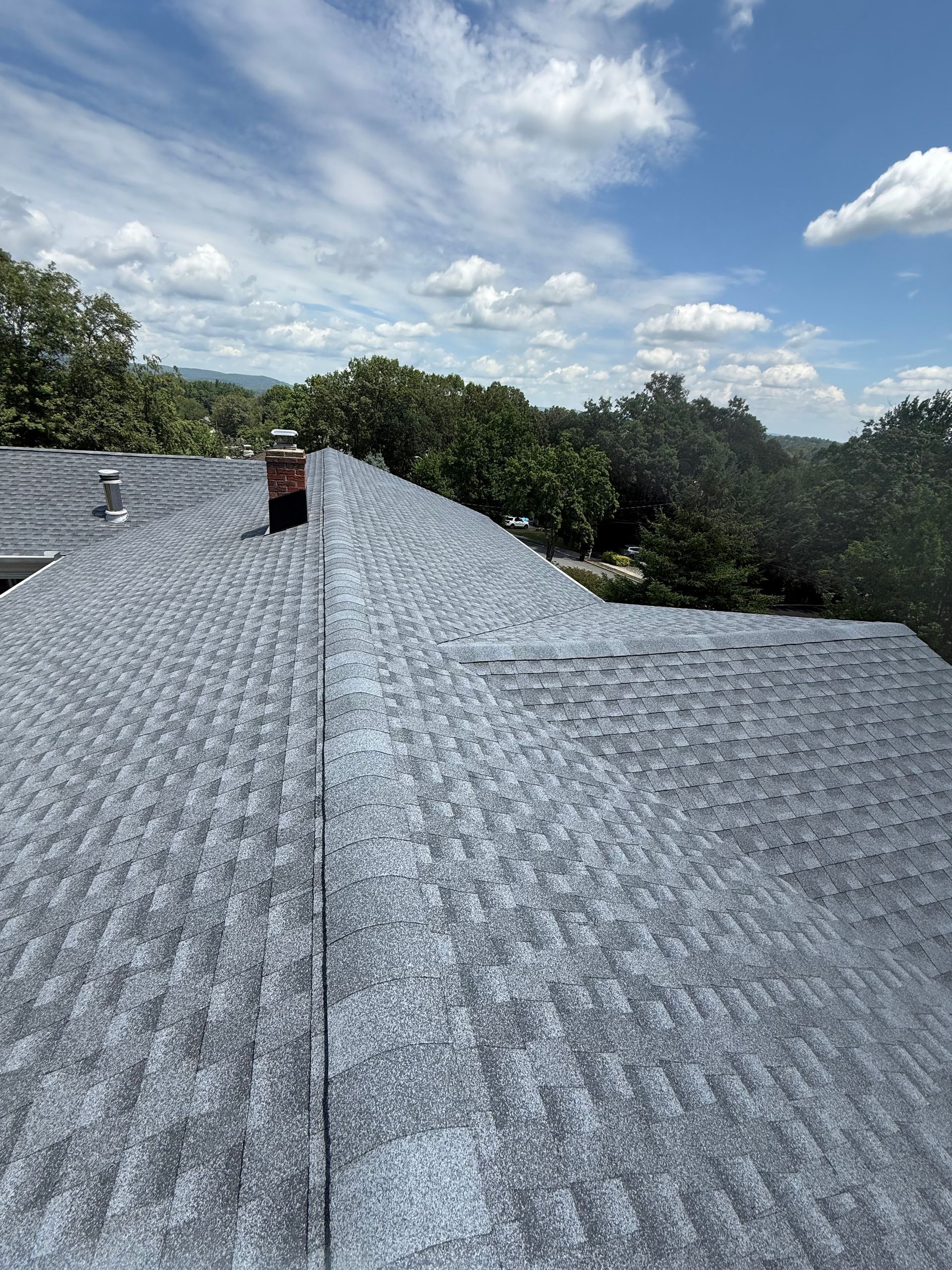 Gray asphalt shingle roof with chimney against a blue sky with fluffy clouds and trees.