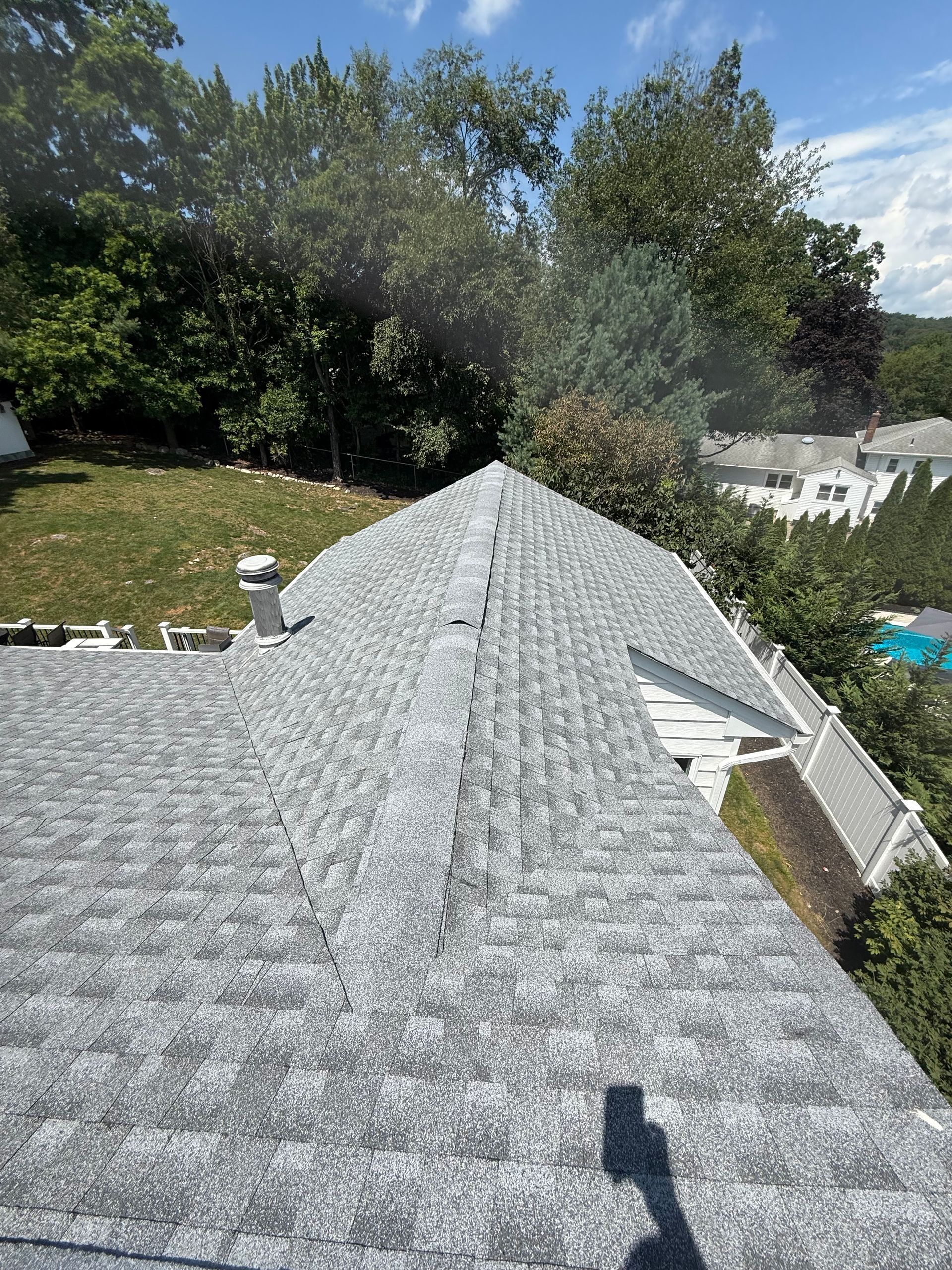 Gray shingled rooftop with a slight slope, surrounded by greenery and a sunny sky.