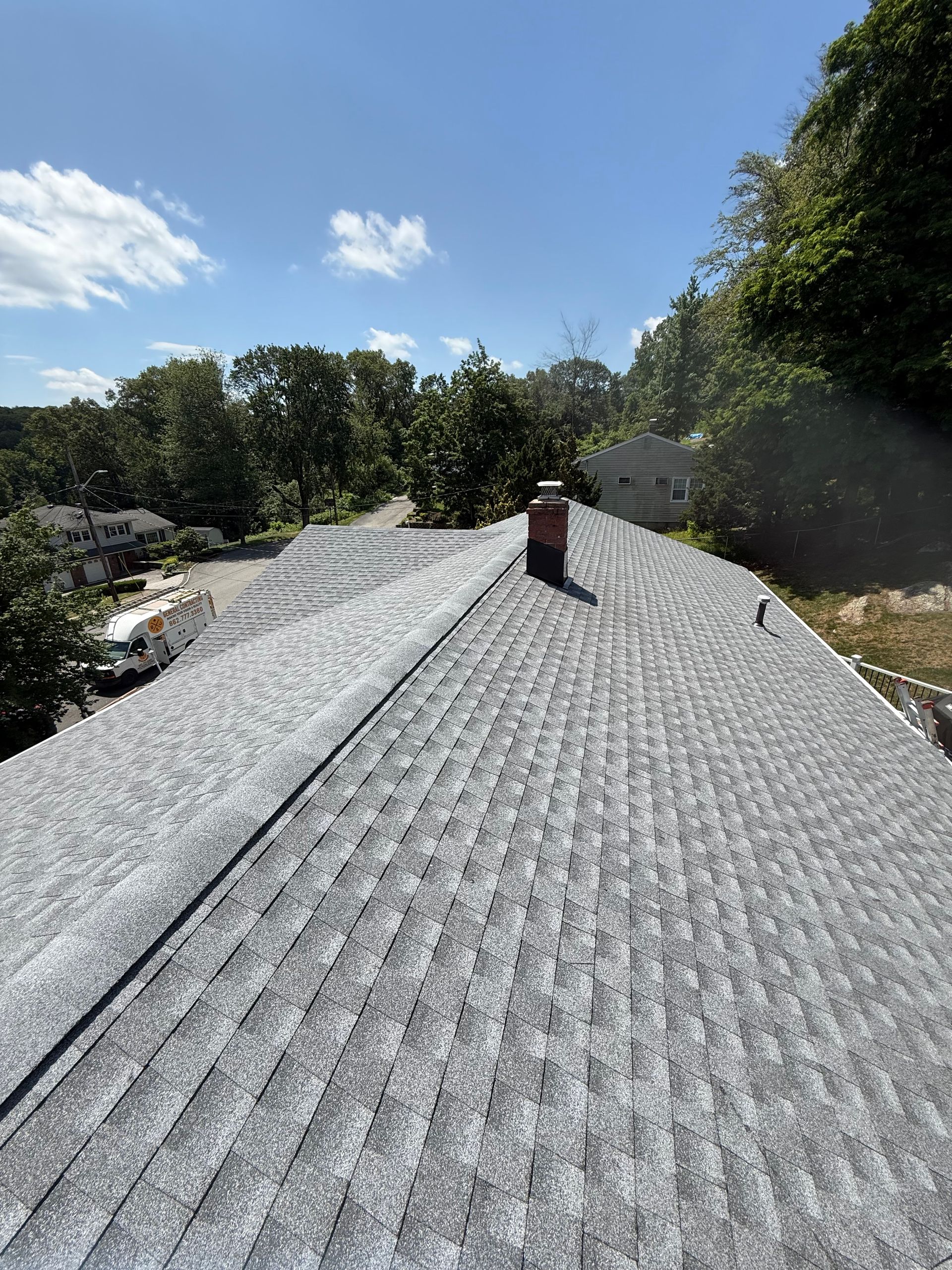 Gray shingle roof of a house with a brick chimney under a bright blue sky. Trees in the background.