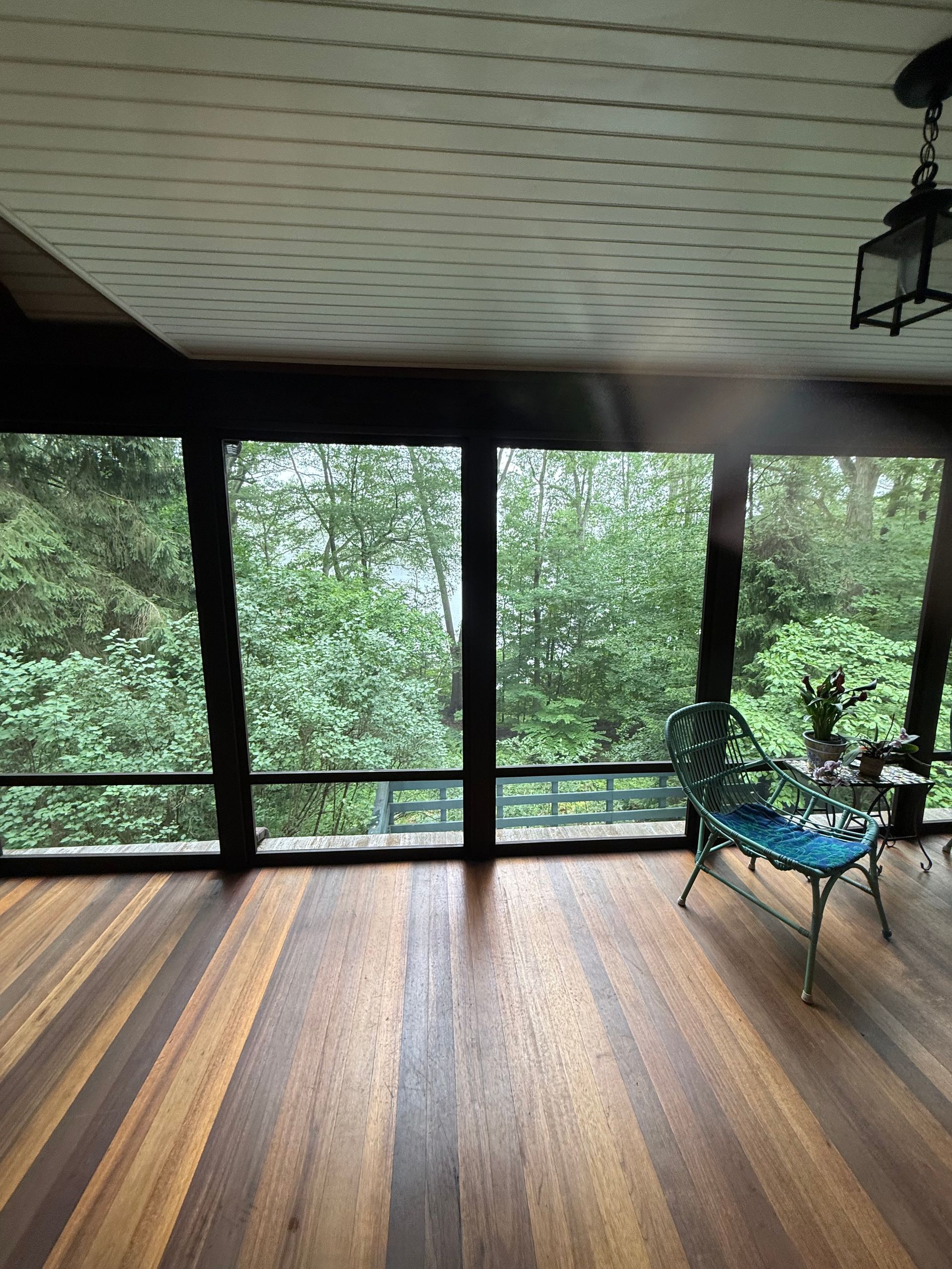 Sunroom with wooden floors, large windows overlooking lush green trees, and a teal bench.