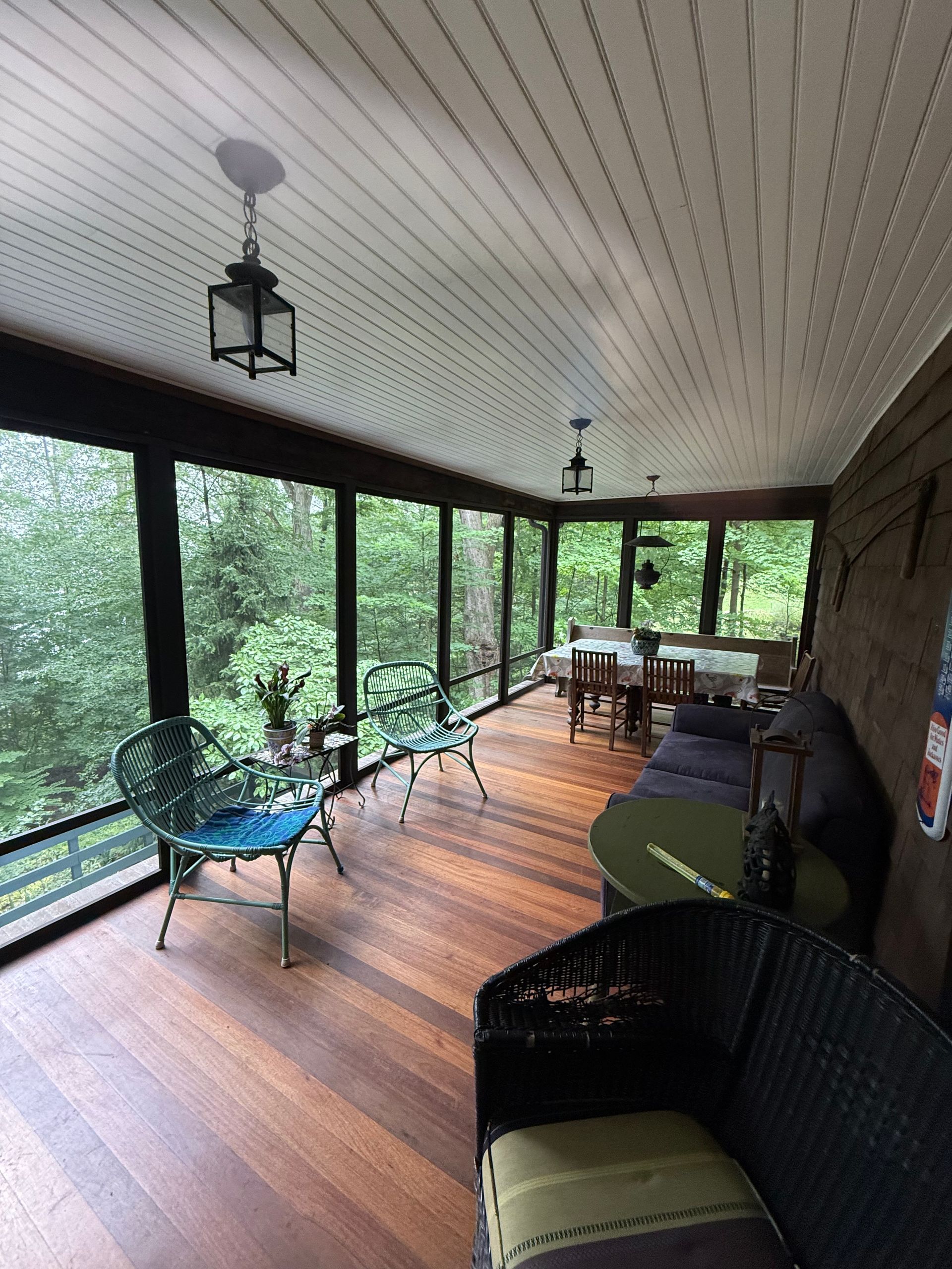 Screened-in porch with wood floor, dark frames, and forest view. Furnished with seating, small tables, and a dining table.