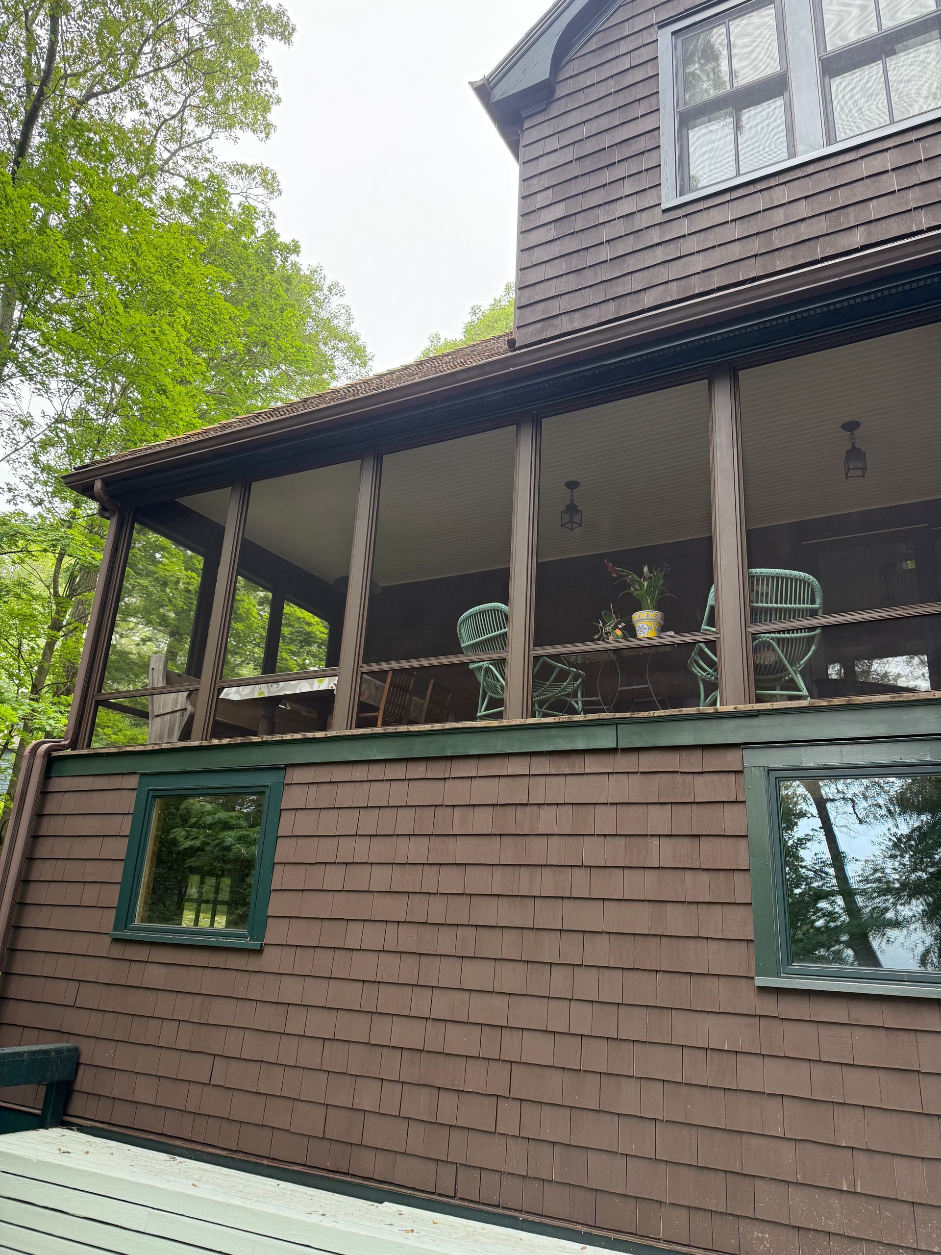 Brown shingled house with porch and two green-framed windows. Trees and overcast sky in the background.