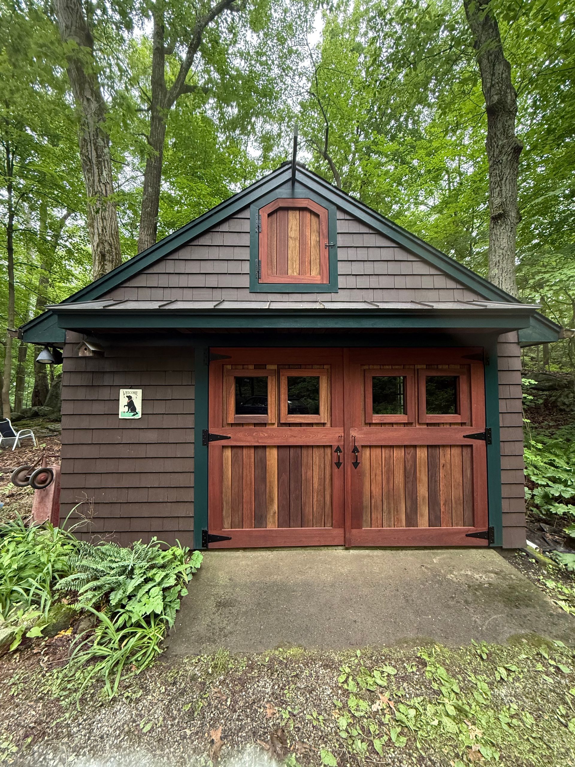 Brown wooden garage with red doors and trim, nestled in a wooded area.