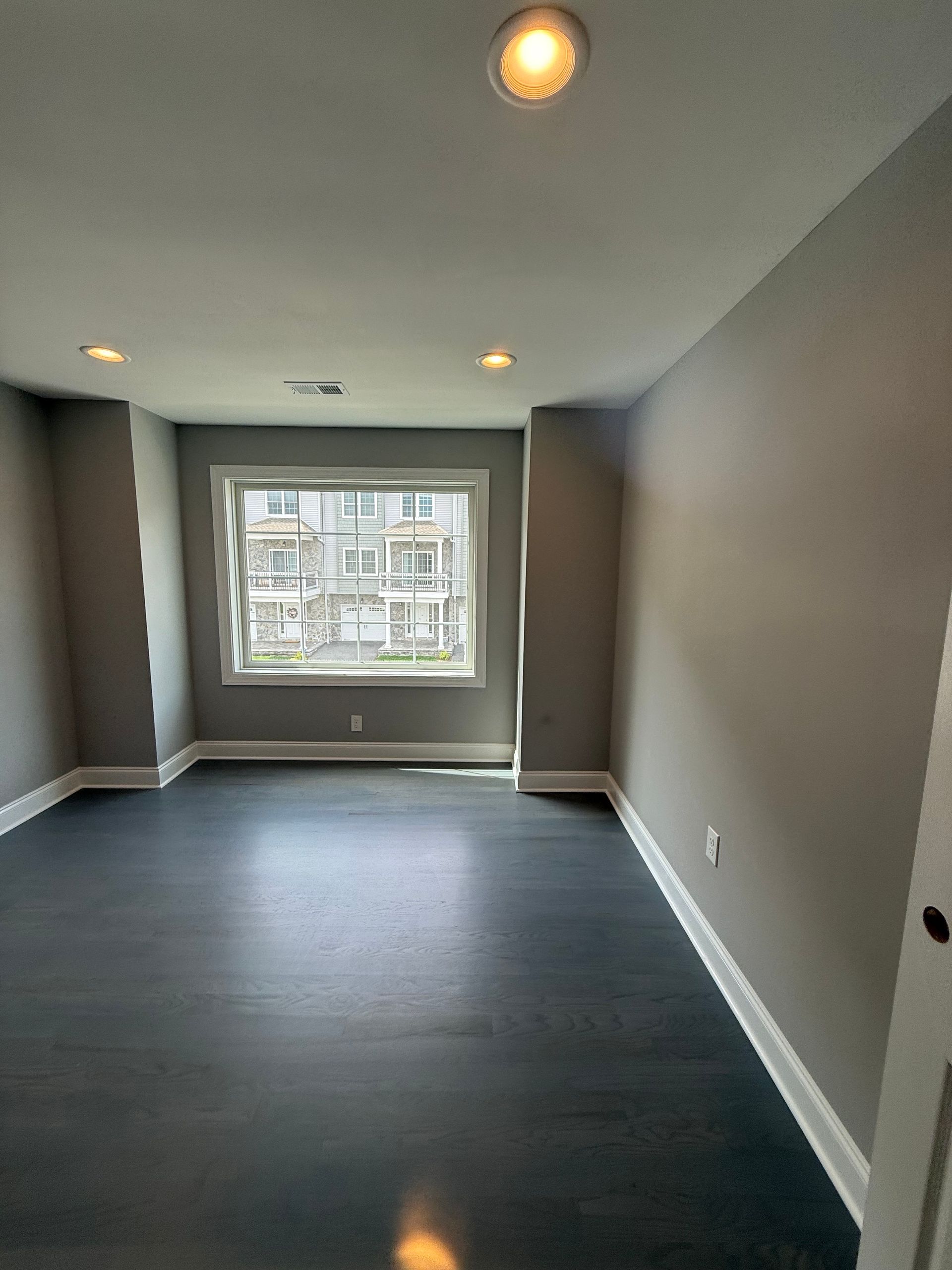 Empty room with dark wood floor, gray walls, white trim, window, and recessed lighting.