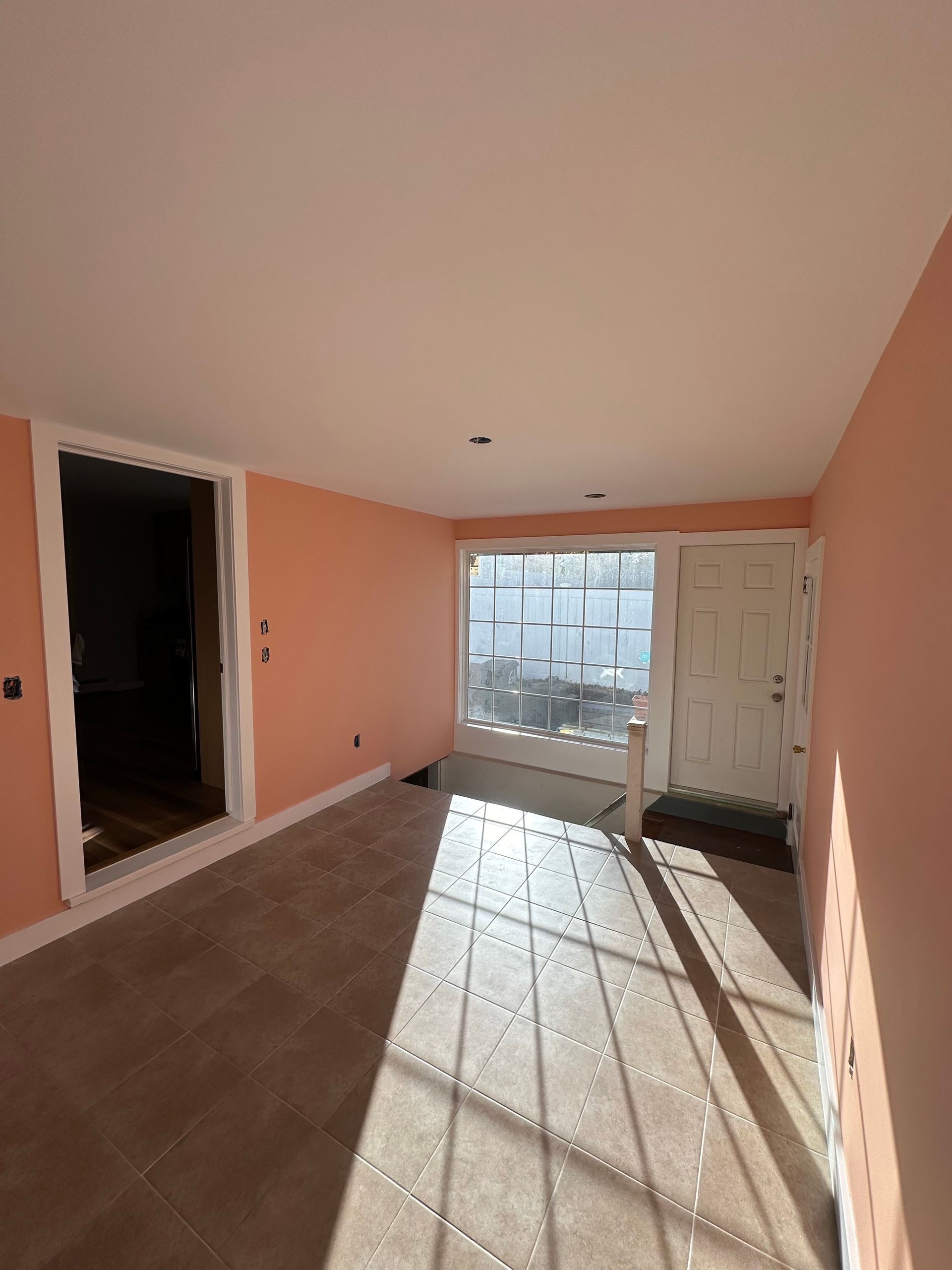 Empty room with peach walls, tiled floor, white door and window letting in bright sunlight.