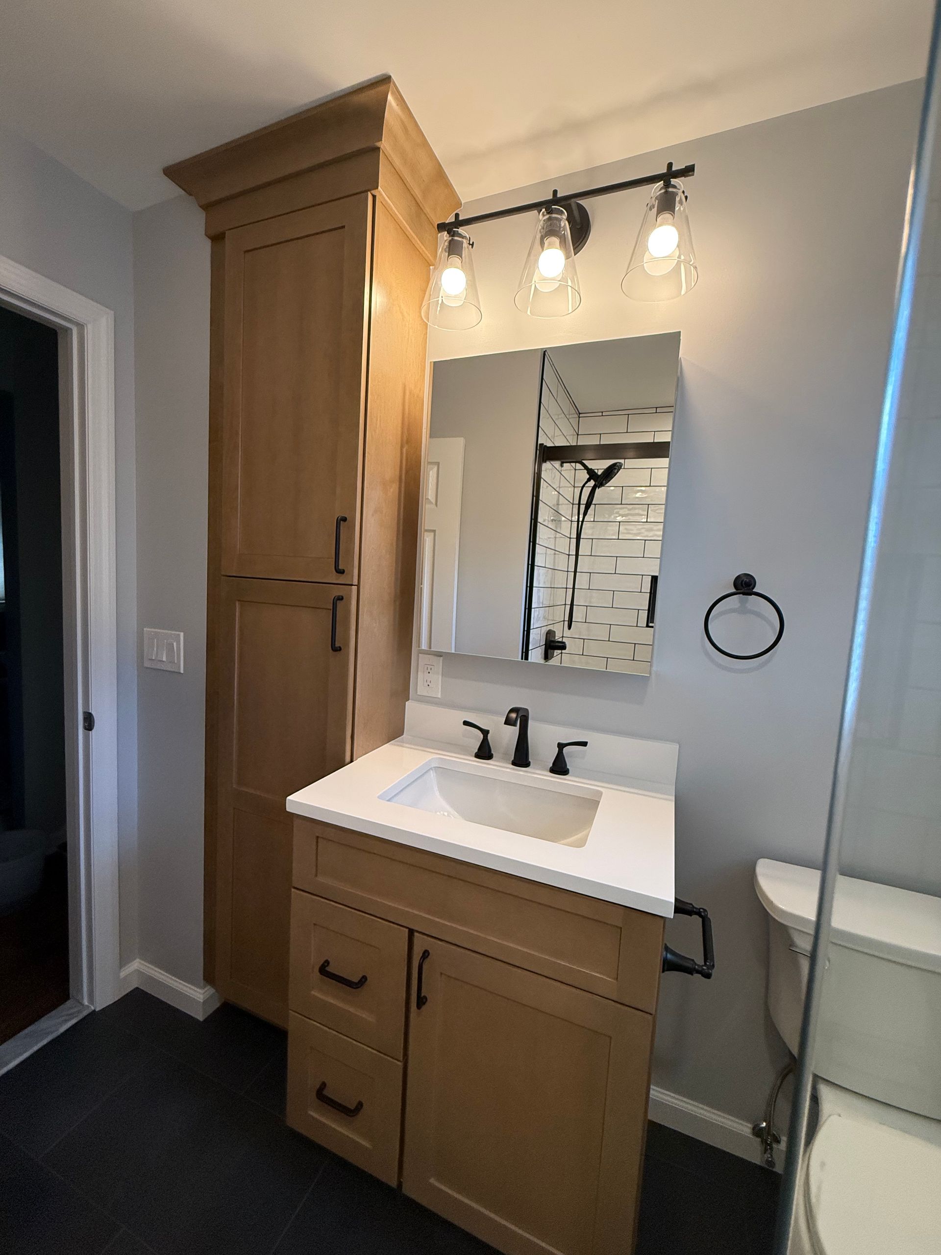 Bathroom with light wood vanity, tall cabinet, sink, mirror, black fixtures, and gray walls.