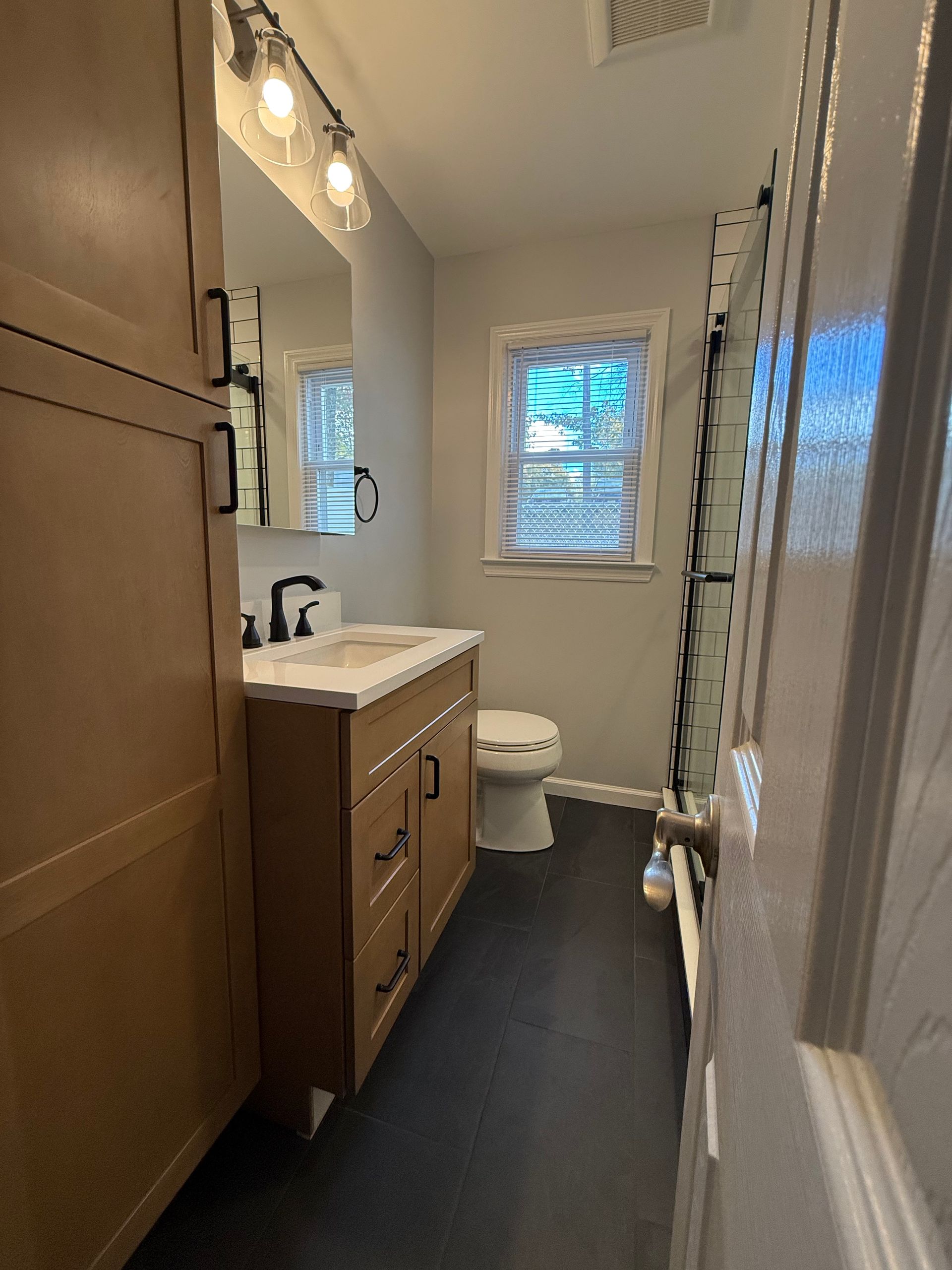 Bathroom with a vanity, toilet, cabinet, and a window with blinds. Dark flooring and light walls.