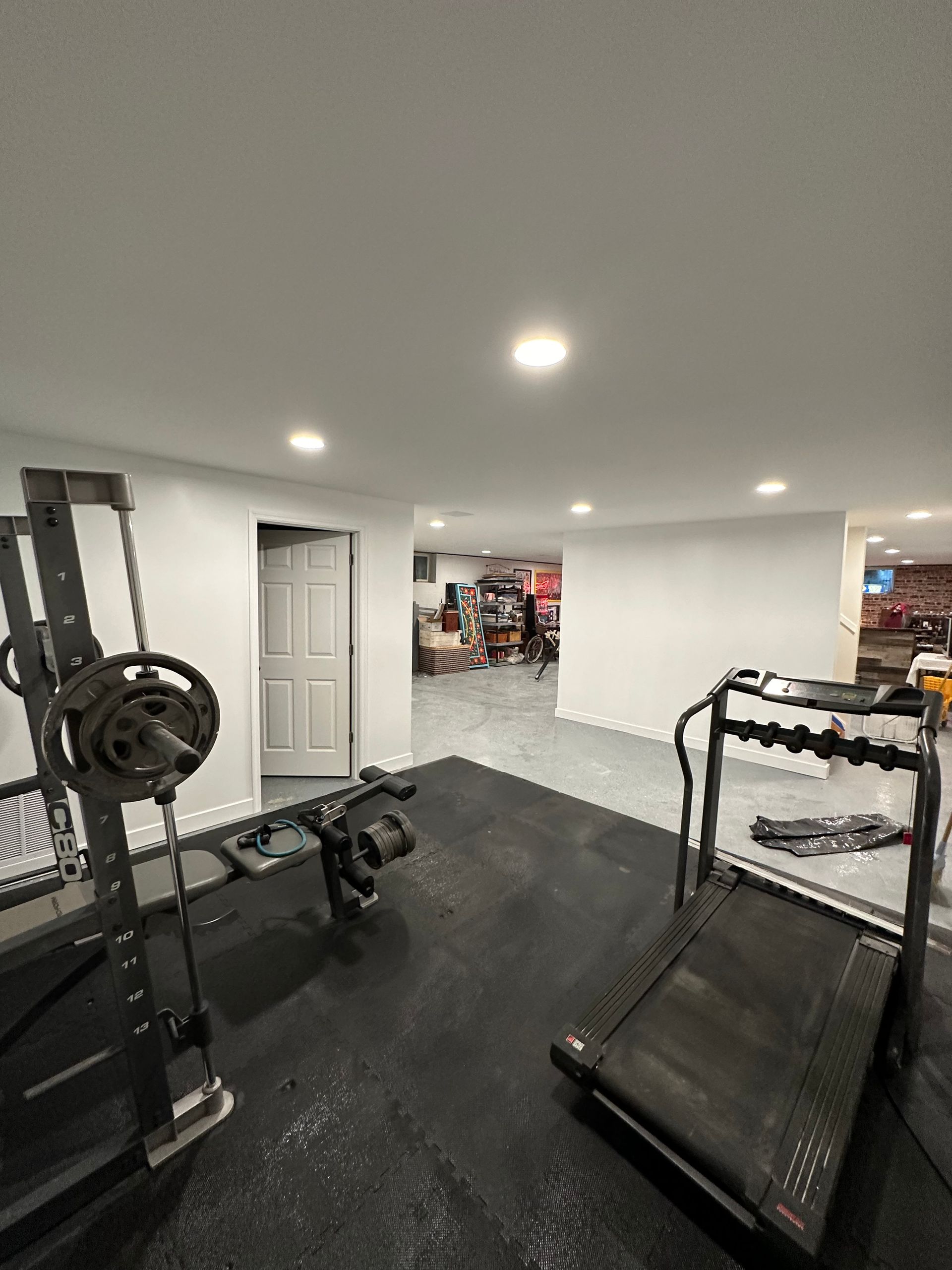 Home gym with weight rack, treadmill, and black rubber flooring. White walls and ceiling, with door.