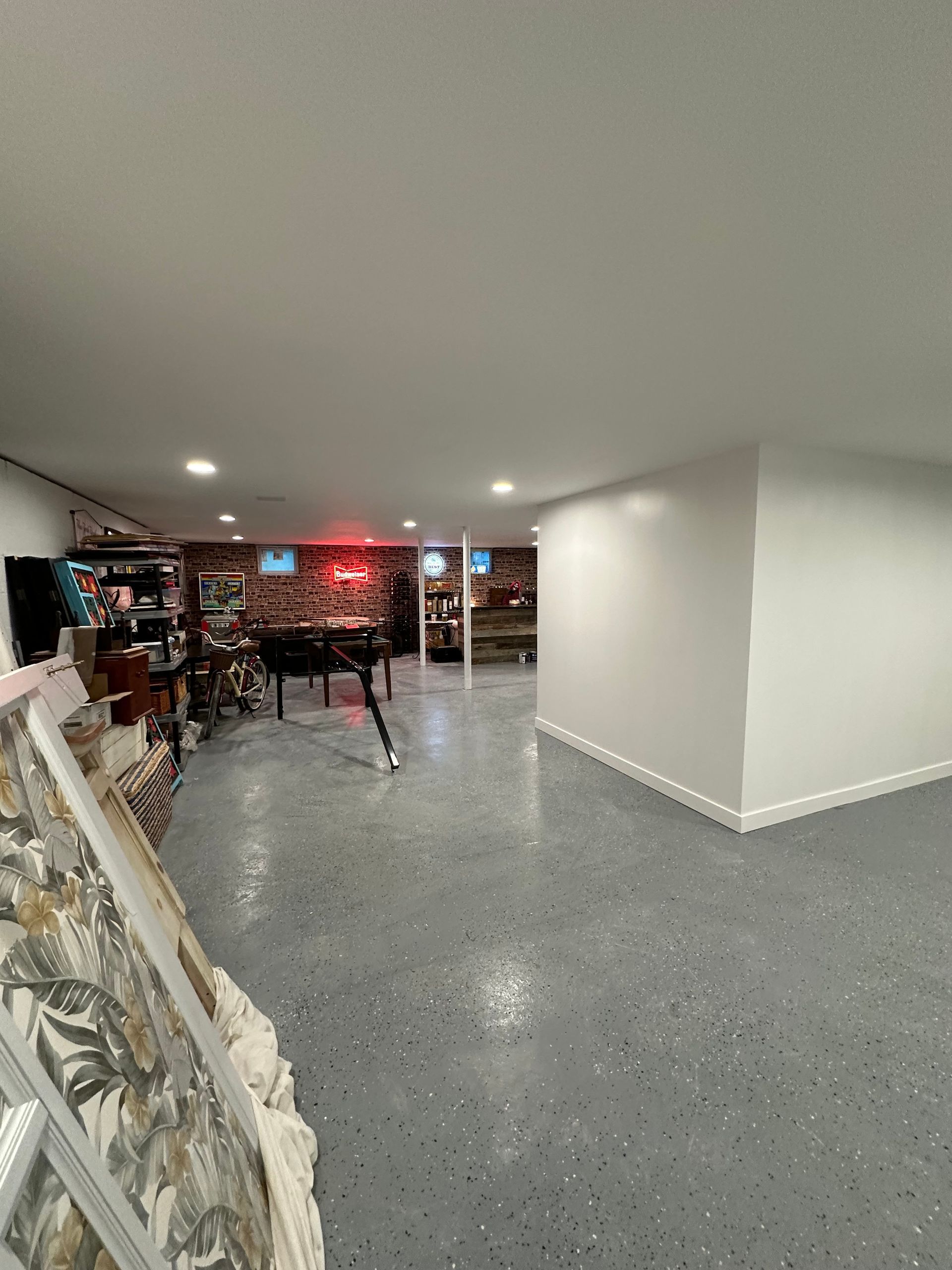 Spacious basement with epoxy floor and white walls. Includes brick feature wall, neon sign, and various items.