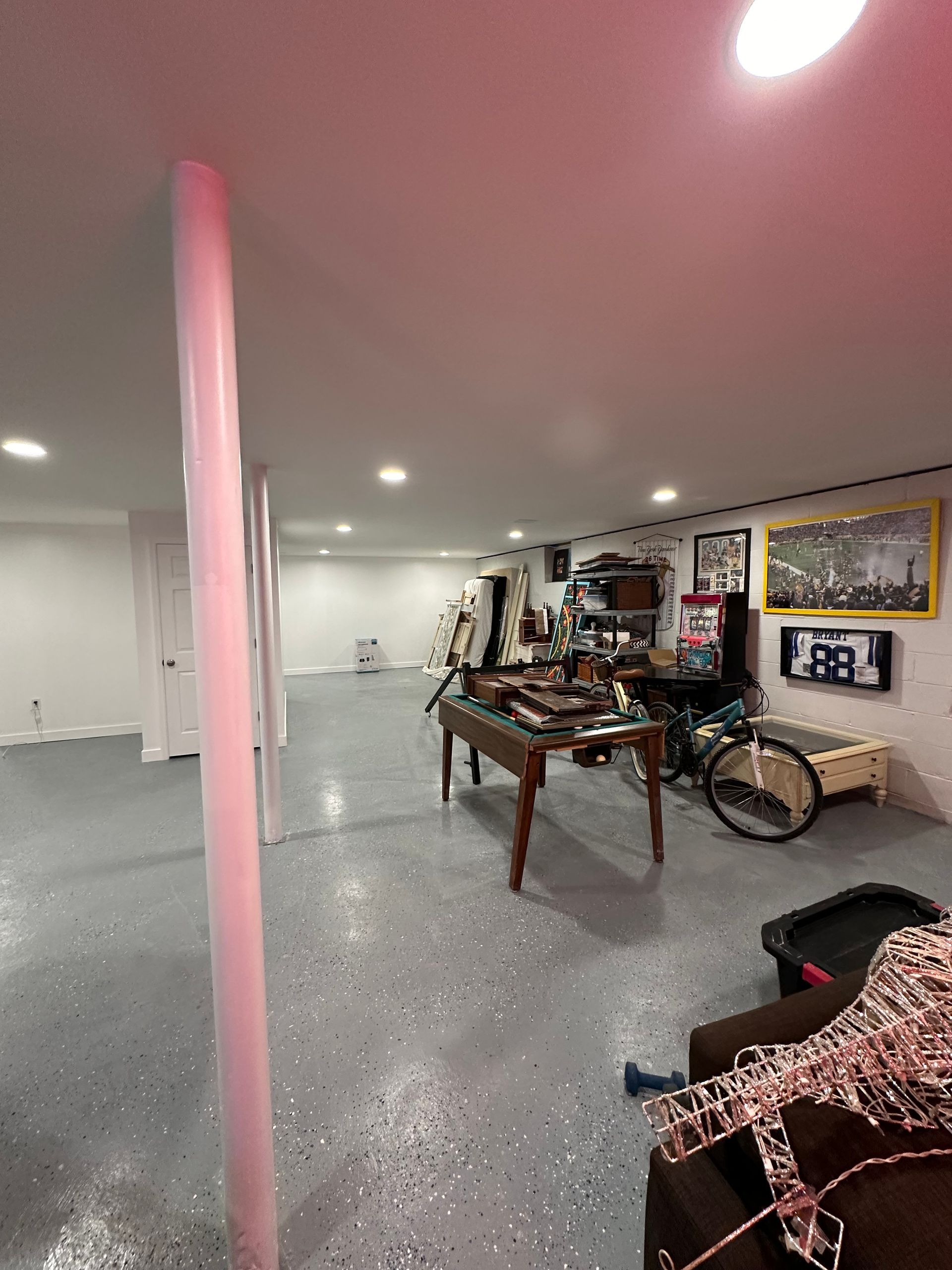 Basement with a support column, various items on a table and against a wall. Concrete flooring.
