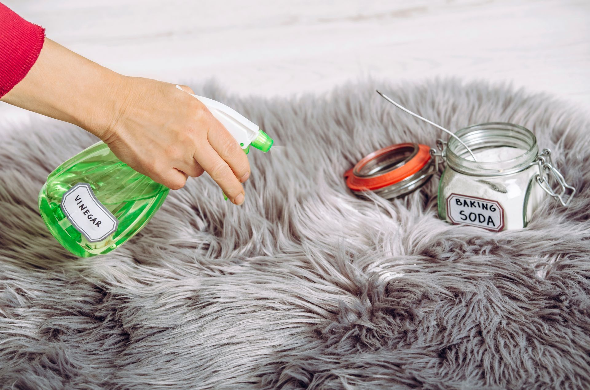 Hand spraying a gray rug with a cleaning solution, next to a jar of baking soda.