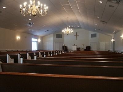 Empty church sanctuary with wooden pews, chandeliers, and a cross on the front wall.