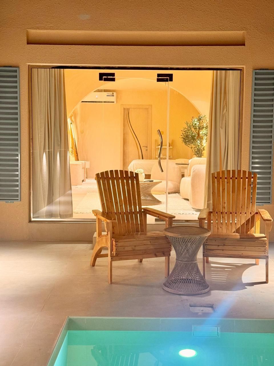 Two wooden chairs and a small table on a patio overlooking a room with white furniture, near a pool.