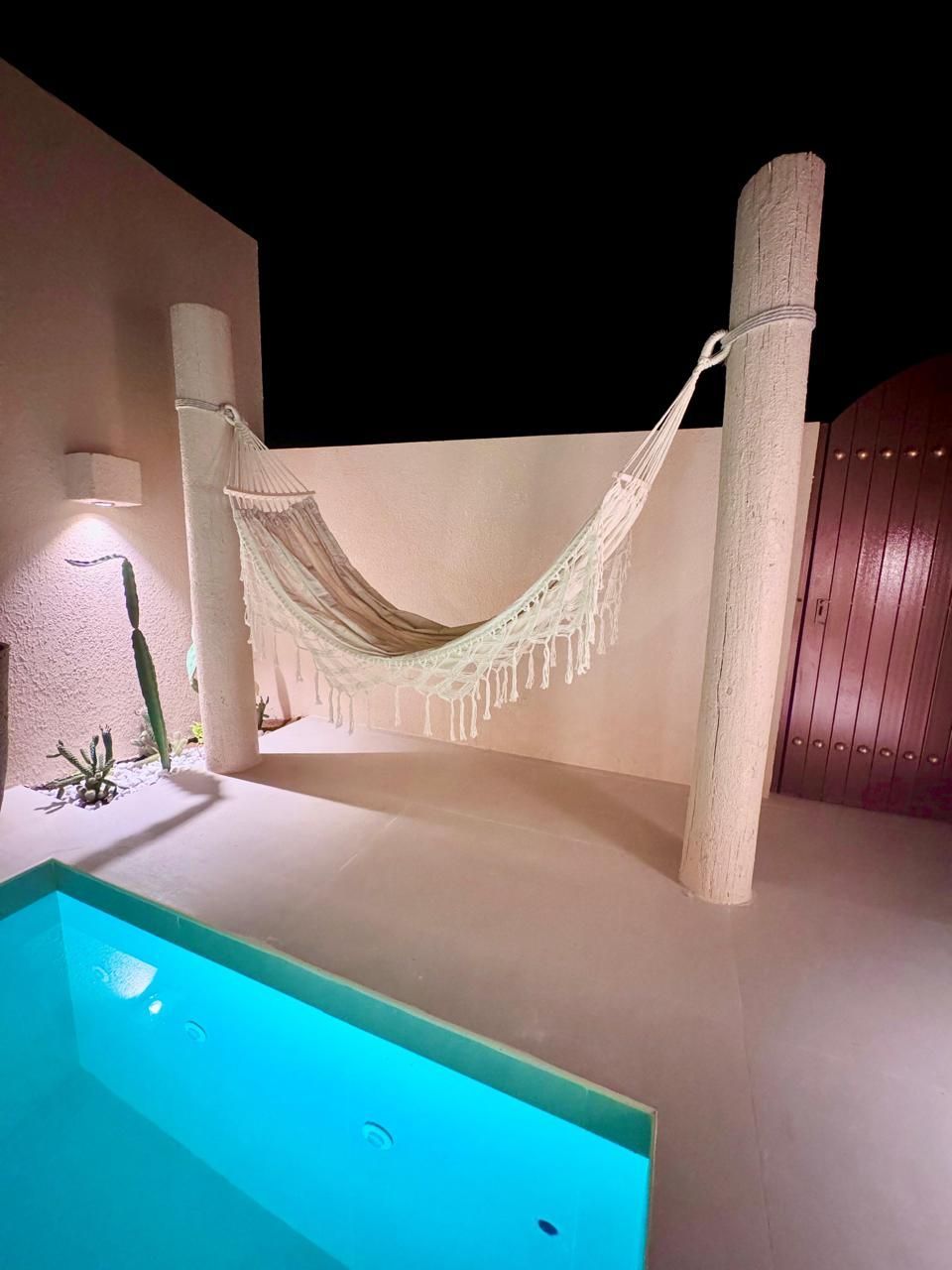 Outdoor scene at night: hammock strung between two white pillars, with a pool in the foreground.