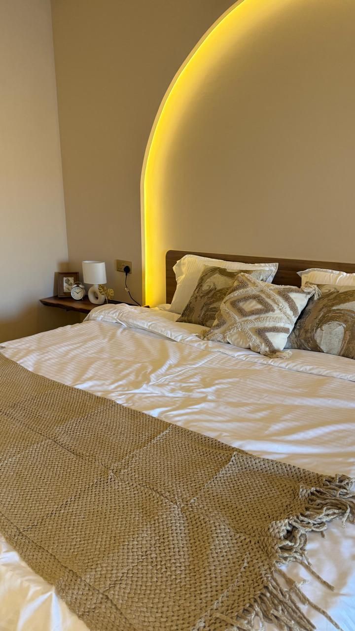 Bedroom with bed, pillows, blanket, and wall-mounted shelf with items. A warm-toned light arch is above the bed.