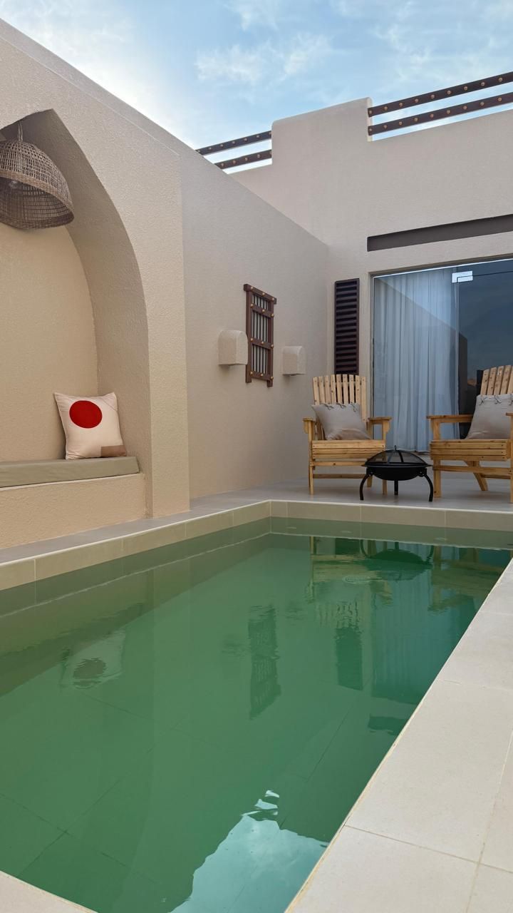 Small outdoor pool with seating, a cushion, and chairs under a light blue sky.