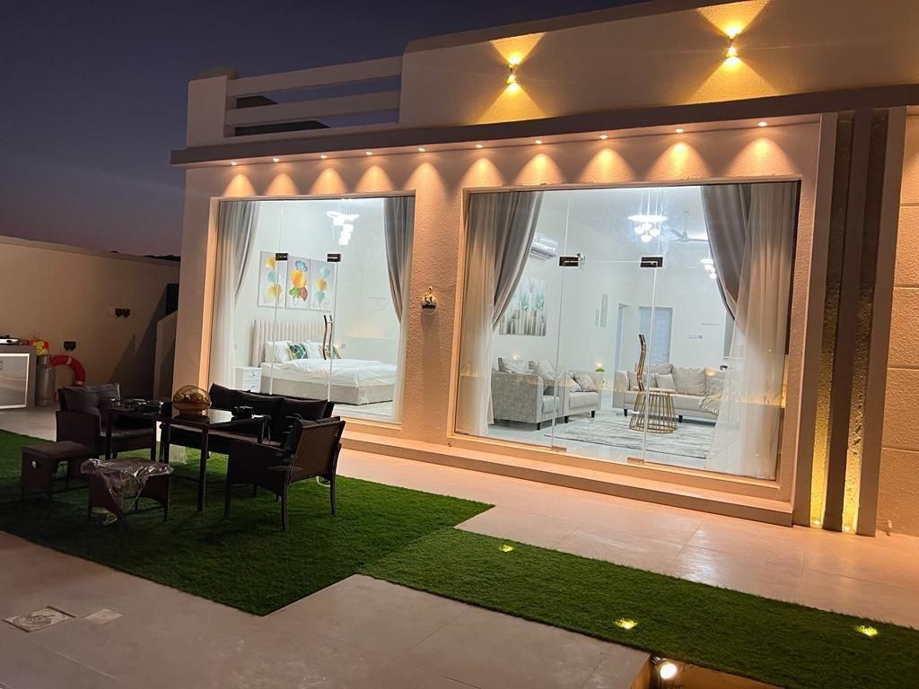 Exterior view of a modern white building at dusk, featuring a seating area with grass and interior lights.