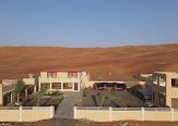 Buildings with brown roofs and beige walls in front of a sand dune under a blue sky.