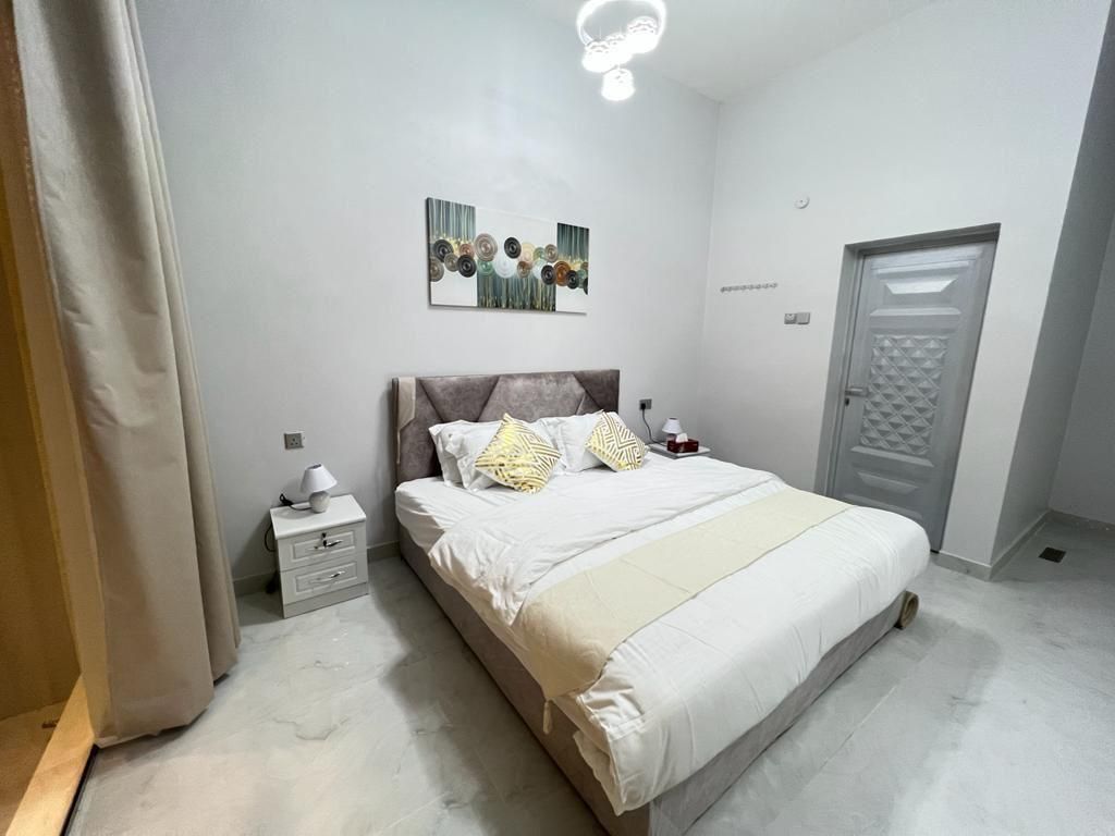 Bedroom with a bed, nightstand, artwork, and a closed gray door. White walls and floor.