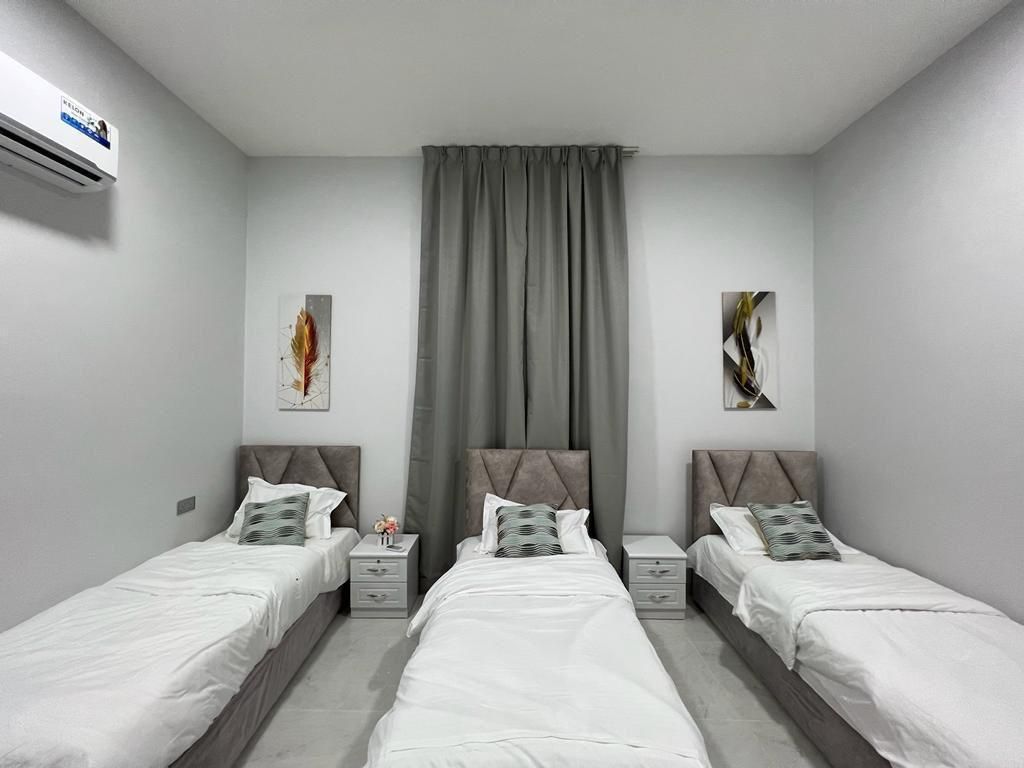 Three single beds in a light gray room with art and a window covered by a gray curtain.